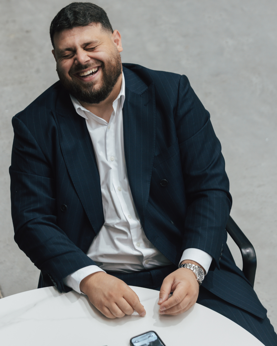 A man in a dark pinstripe suit and white shirt laughing, sitting at a table with a smartphone.