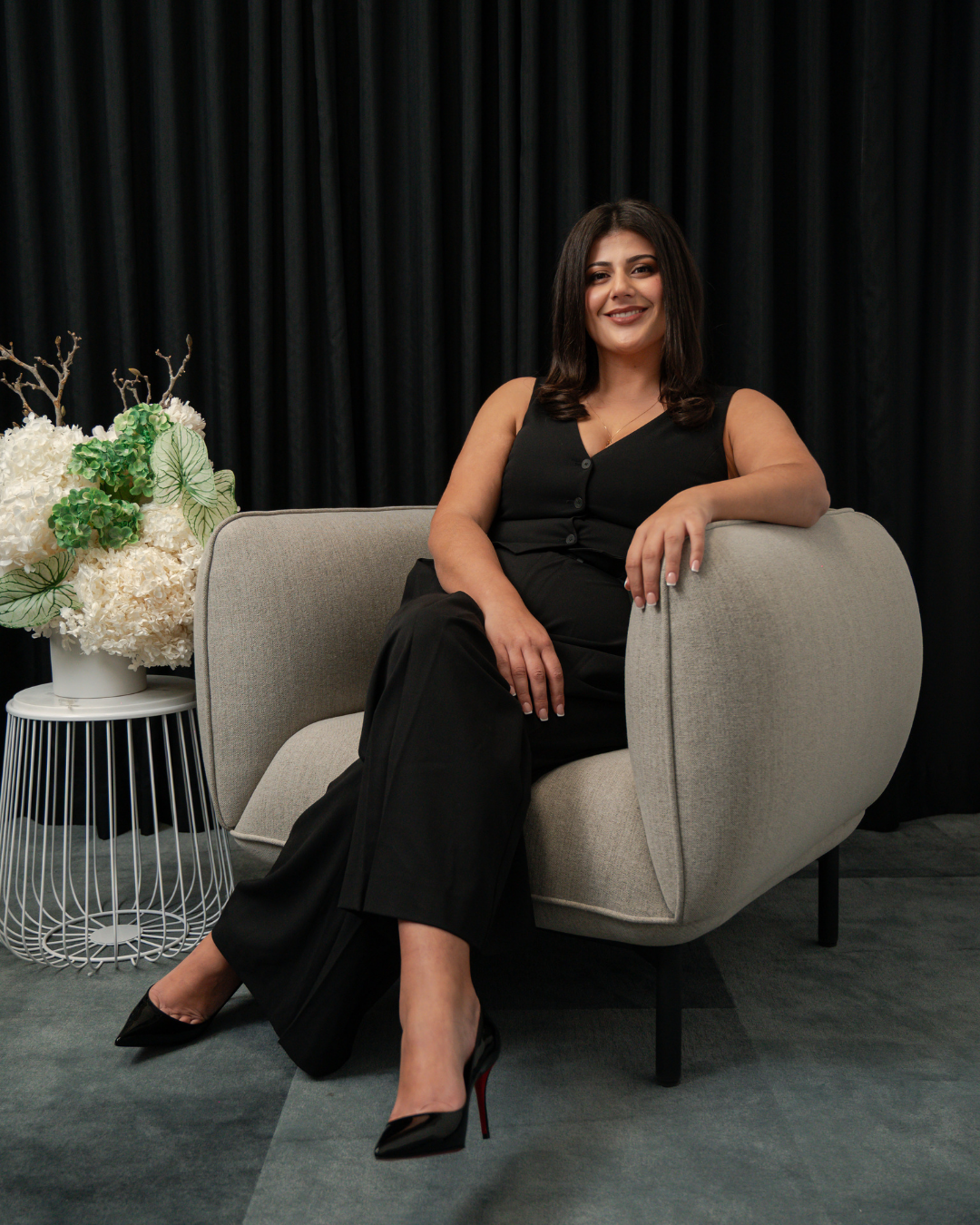Woman wearing black sleeveless outfit and heels sitting on a modern beige armchair with black curtain backdrop and white floral arrangement on a small side table.