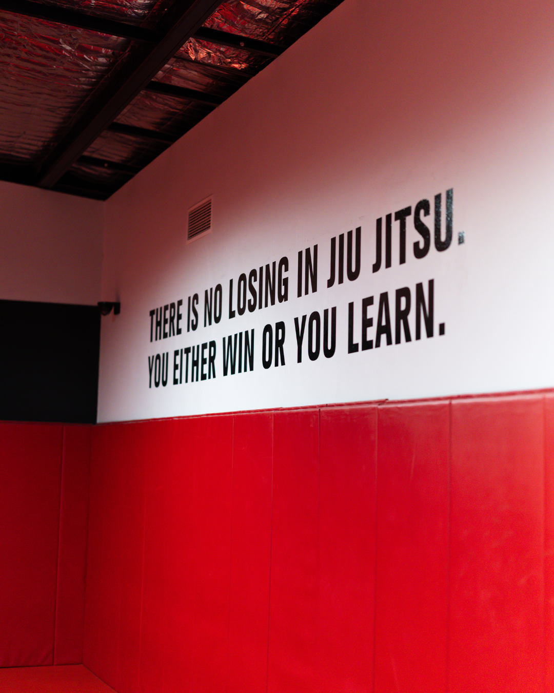 Quote on wall: 'There is no losing in jiu jitsu. You either win or you learn.' The wall is white with black text and part of the room has red padding, likely in a martial arts gym or training center.