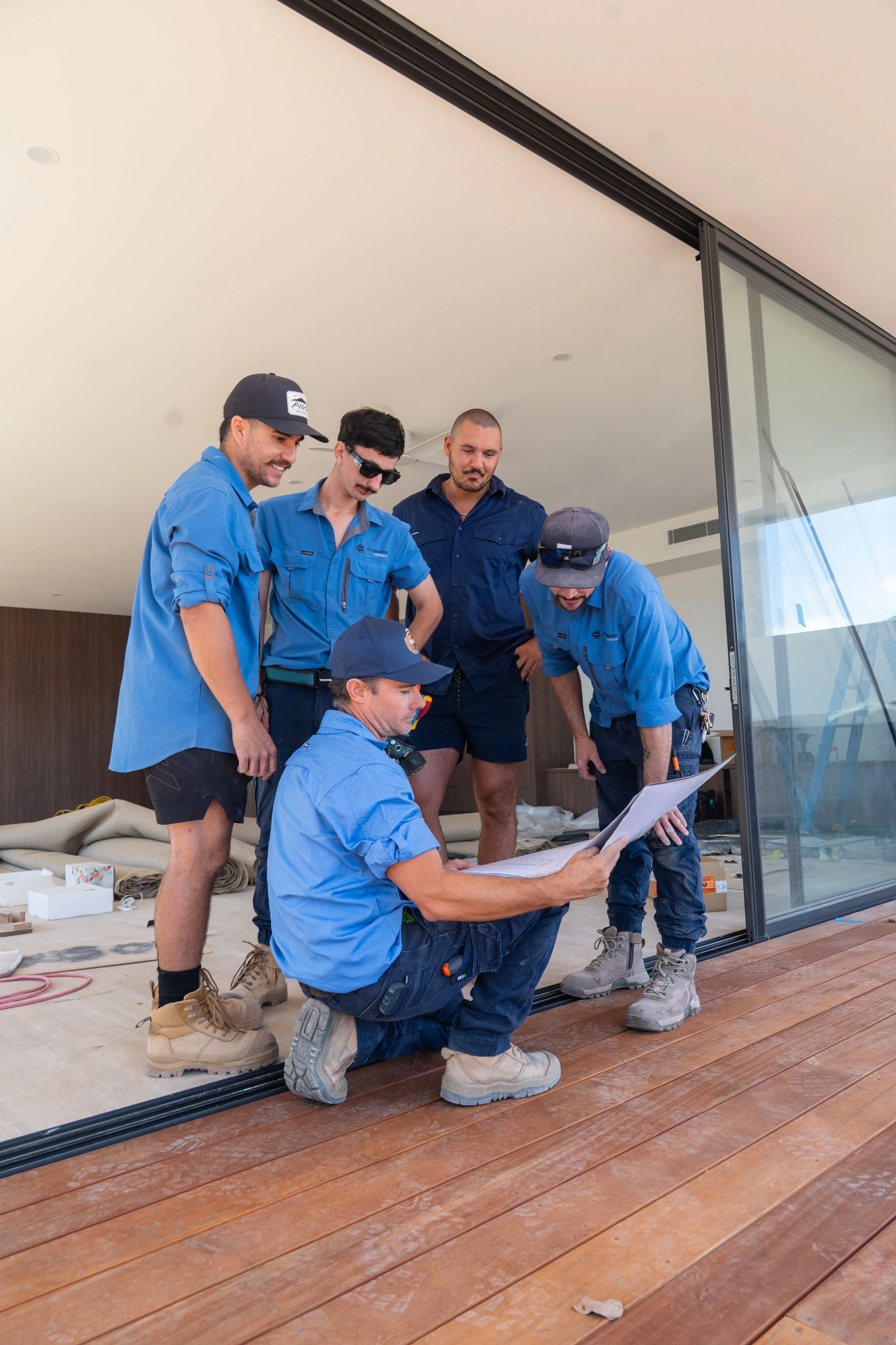 Group of five construction workers, three standing and one sitting, looking at a blueprint inside a modern building. All are wearing blue work shirts and boots.