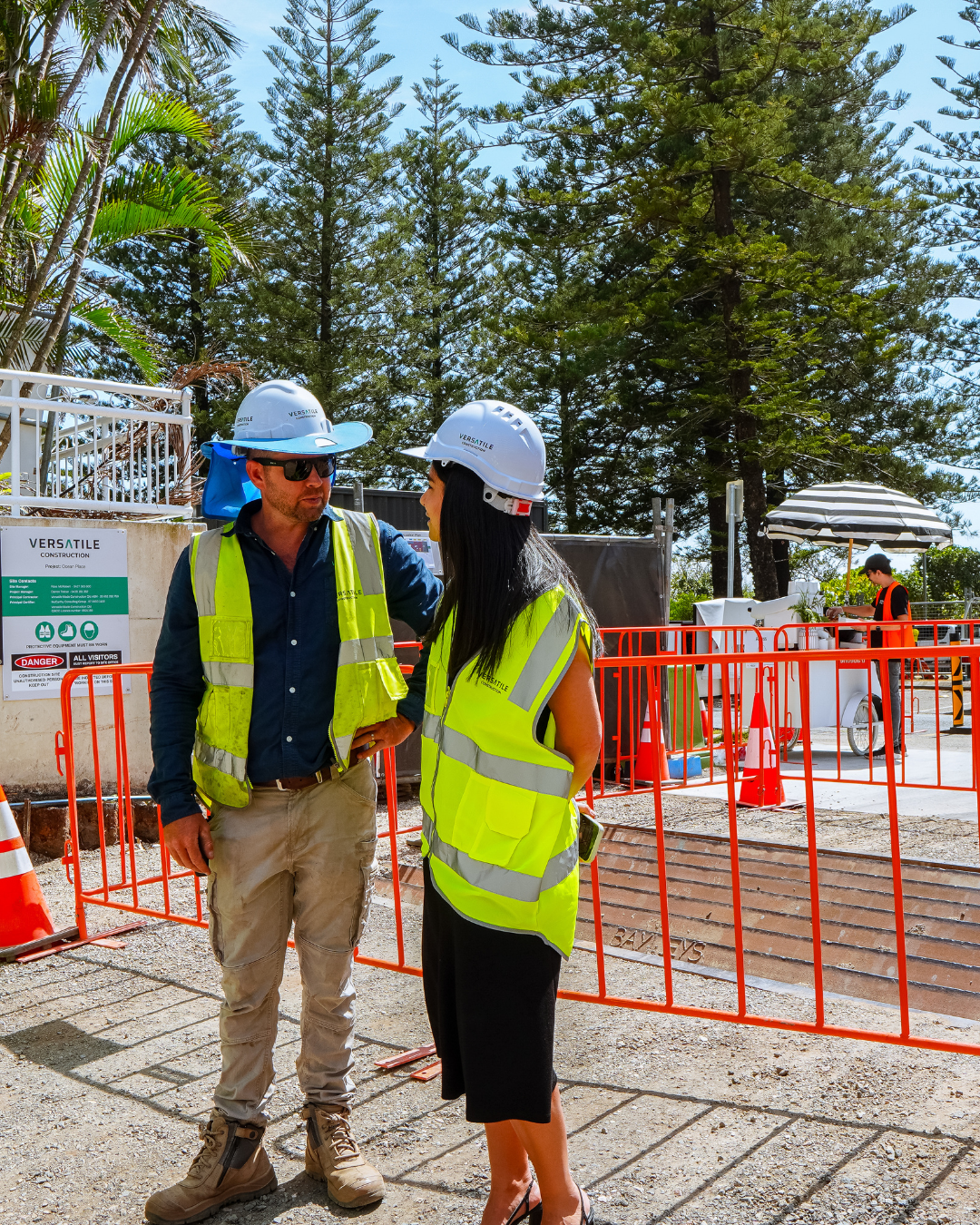 Two construction workers, a man and a woman, are having a conversation at a construction site. They are wearing hard hats, safety vests, and protective gear. Behind them, there are trees, construction signage, and workers, one of whom is under a stri