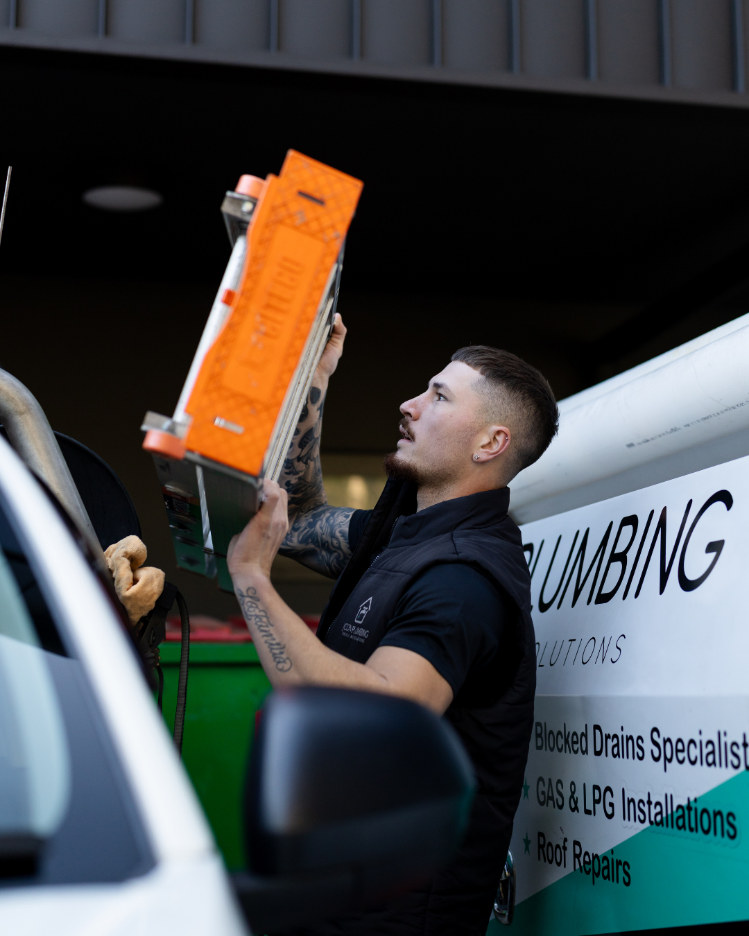 A man with tattoos and a short haircut, wearing a black shirt, is holding and inspecting an orange safety device or ladder near a truck with plumbing services advertising.