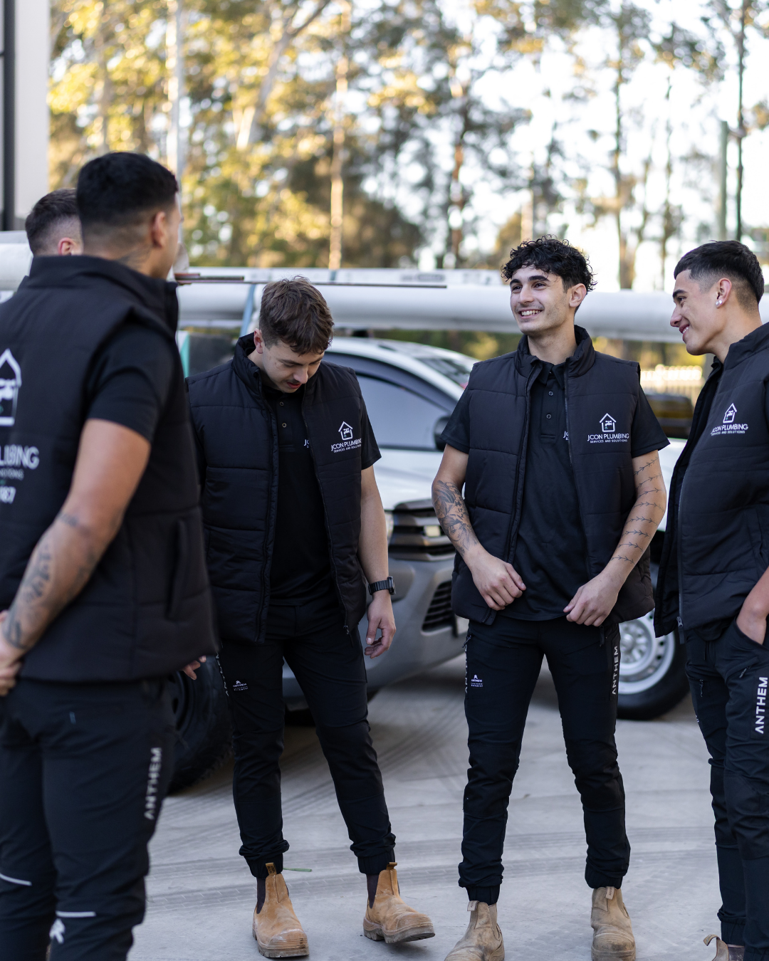 Group of five male workers standing outdoors near a truck, wearing black uniforms with logo, smiling and engaged in conversation, with trees in the background.