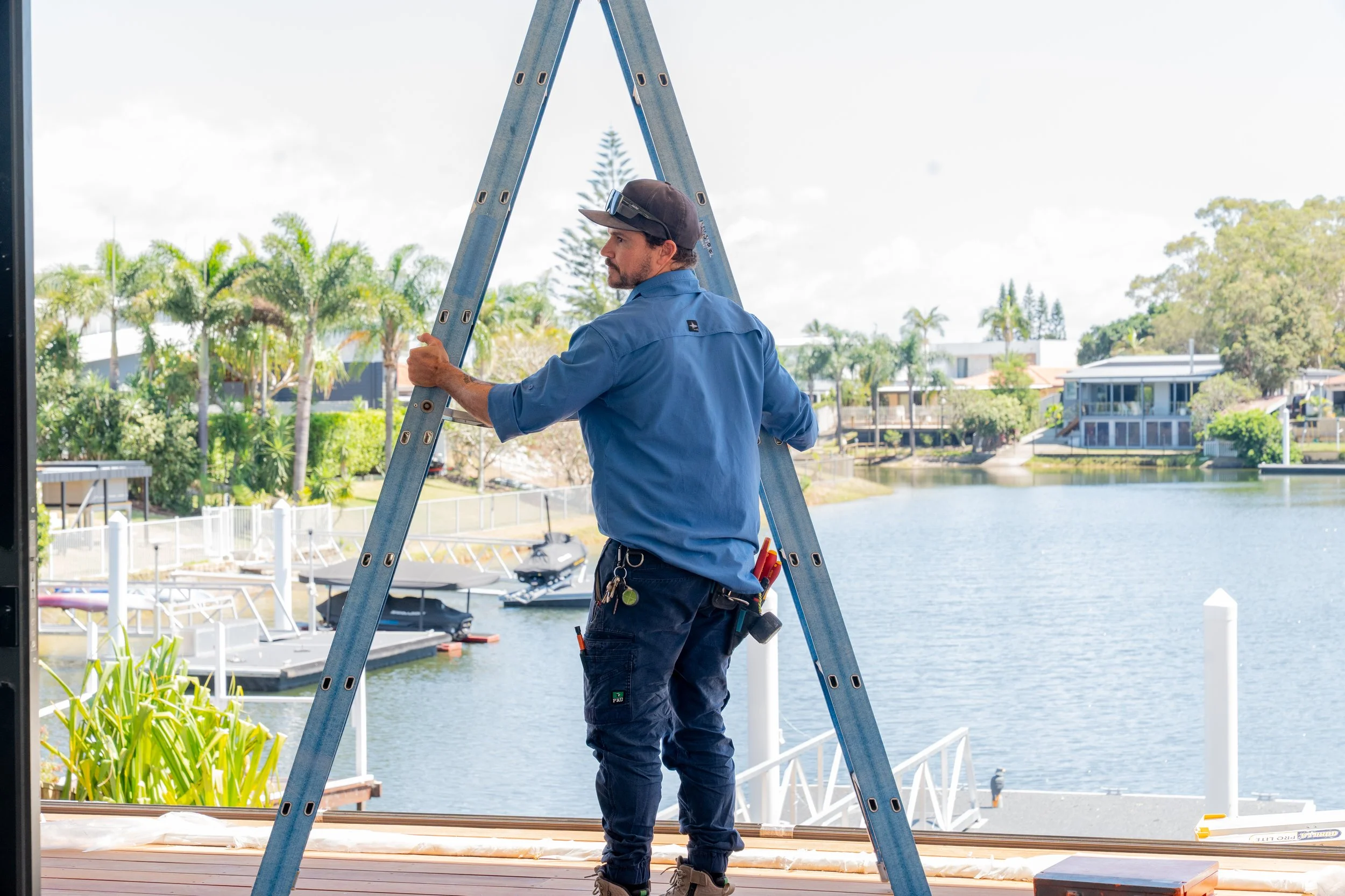 A man in a blue work shirt and dark pants standing on a ladder by a waterfront, with a scenic view of water, boats, trees, and modern houses in the background.