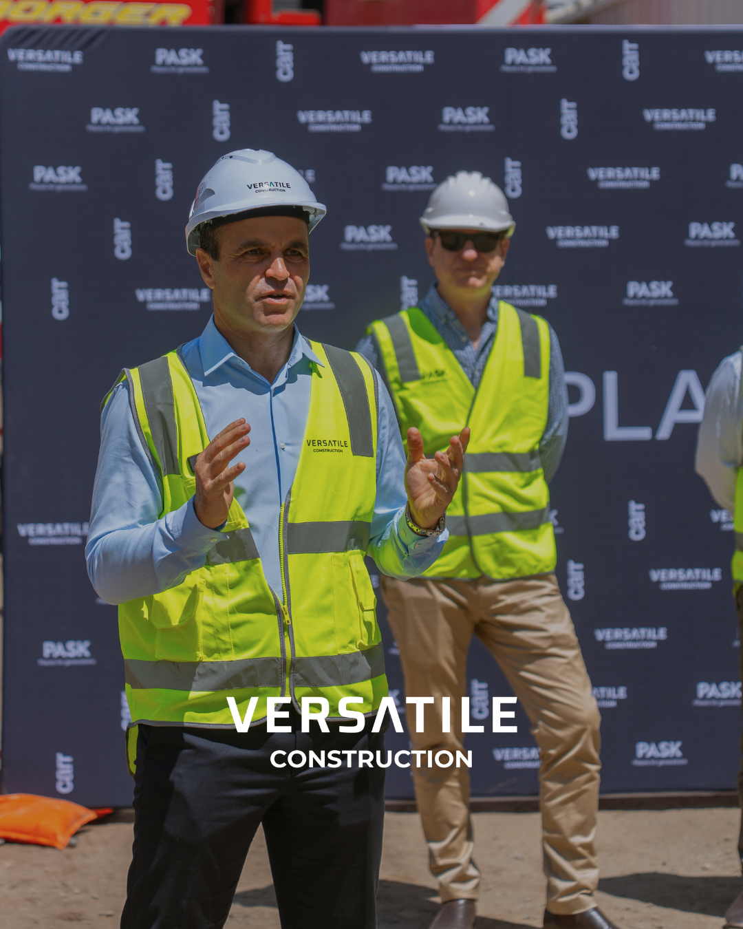 Two men wearing white hard hats and yellow safety vests speaking at a construction site with a black backdrop displaying logos for "Pask" and "Versatile Construction."