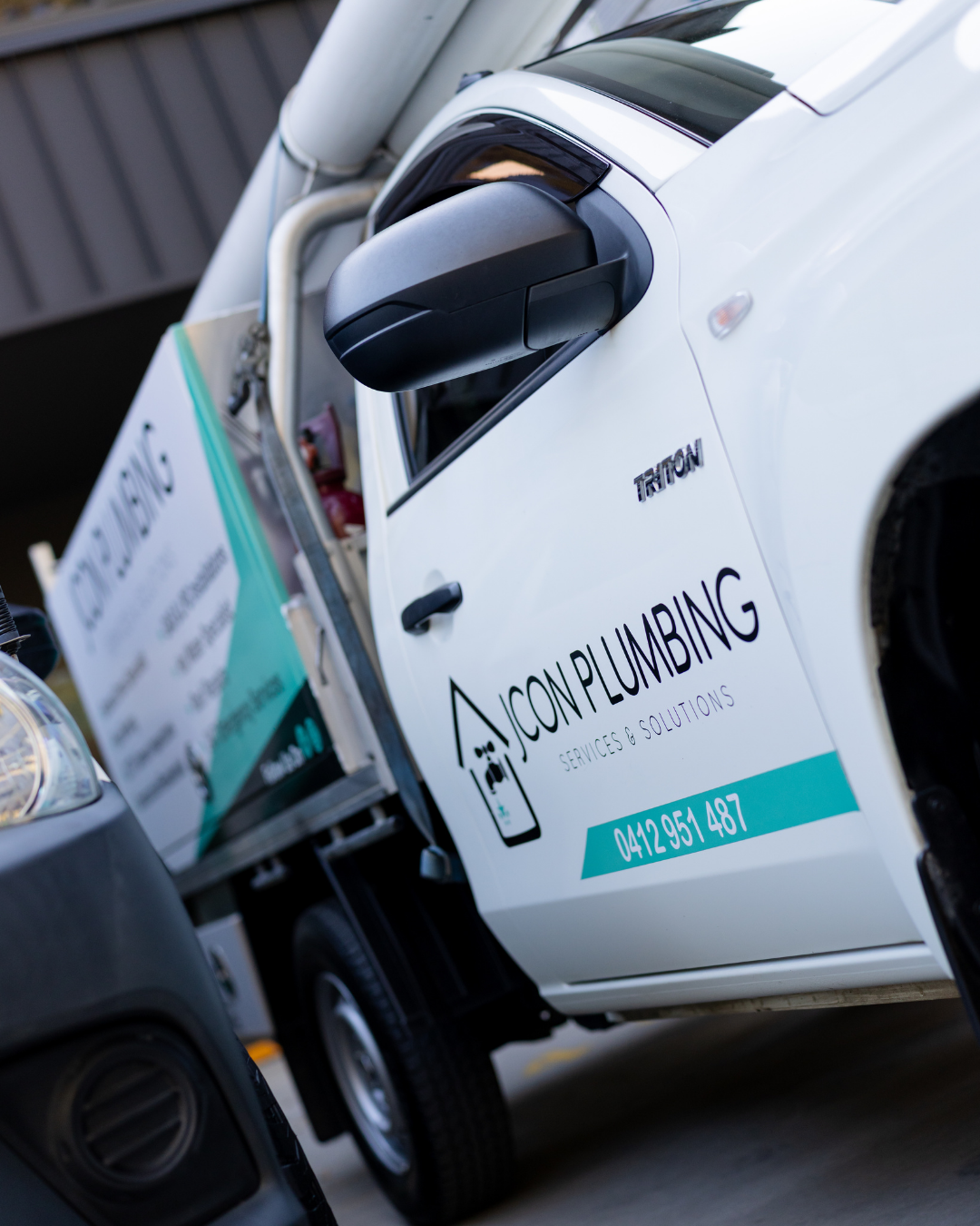 Side view of a white company van with signage for Icon Plumbing, Services & Solutions, and a phone number. Part of another vehicle is visible in the foreground.