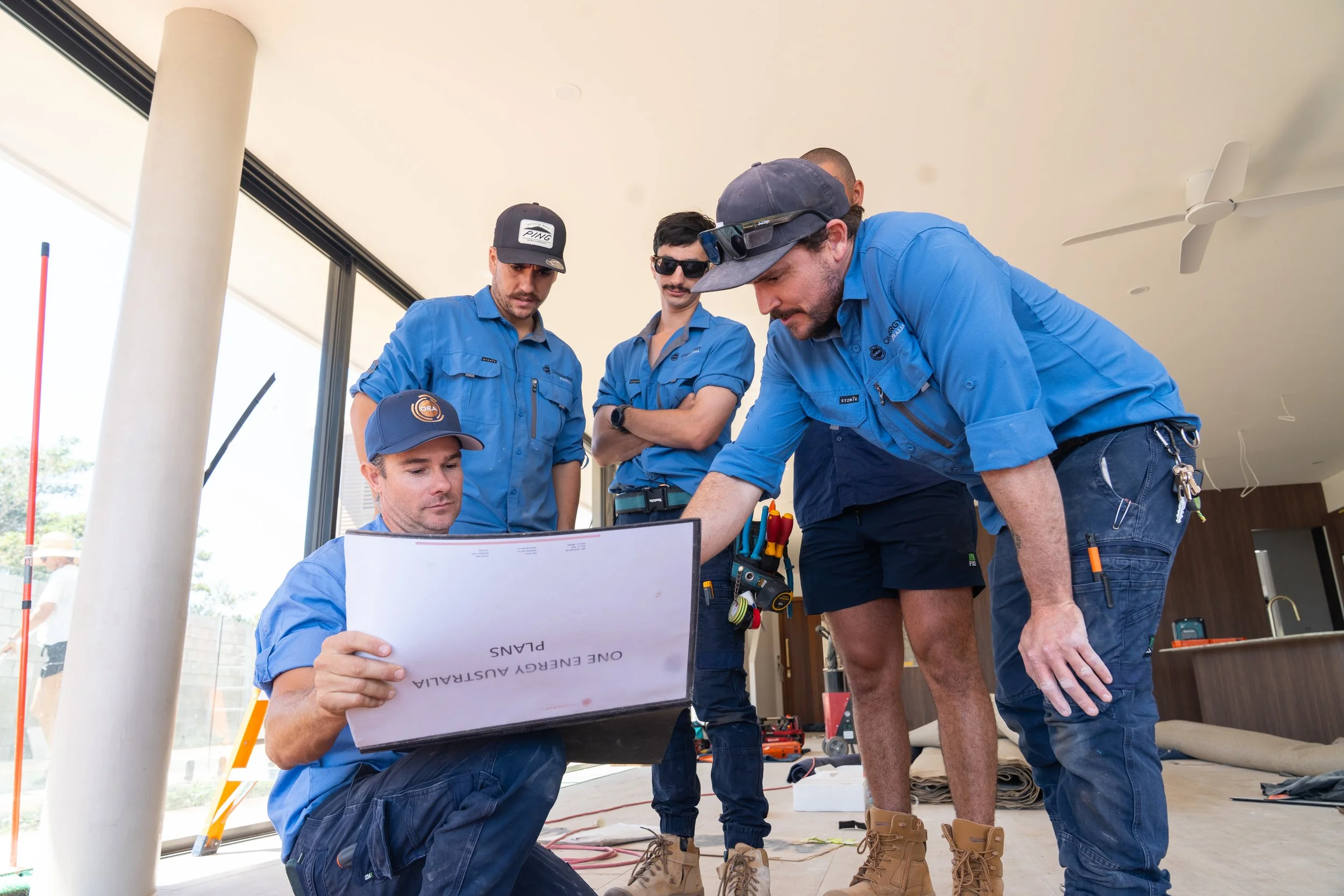 Group of five construction workers in blue shirts gathered around a laptop or blueprint inside a building, discussing something.