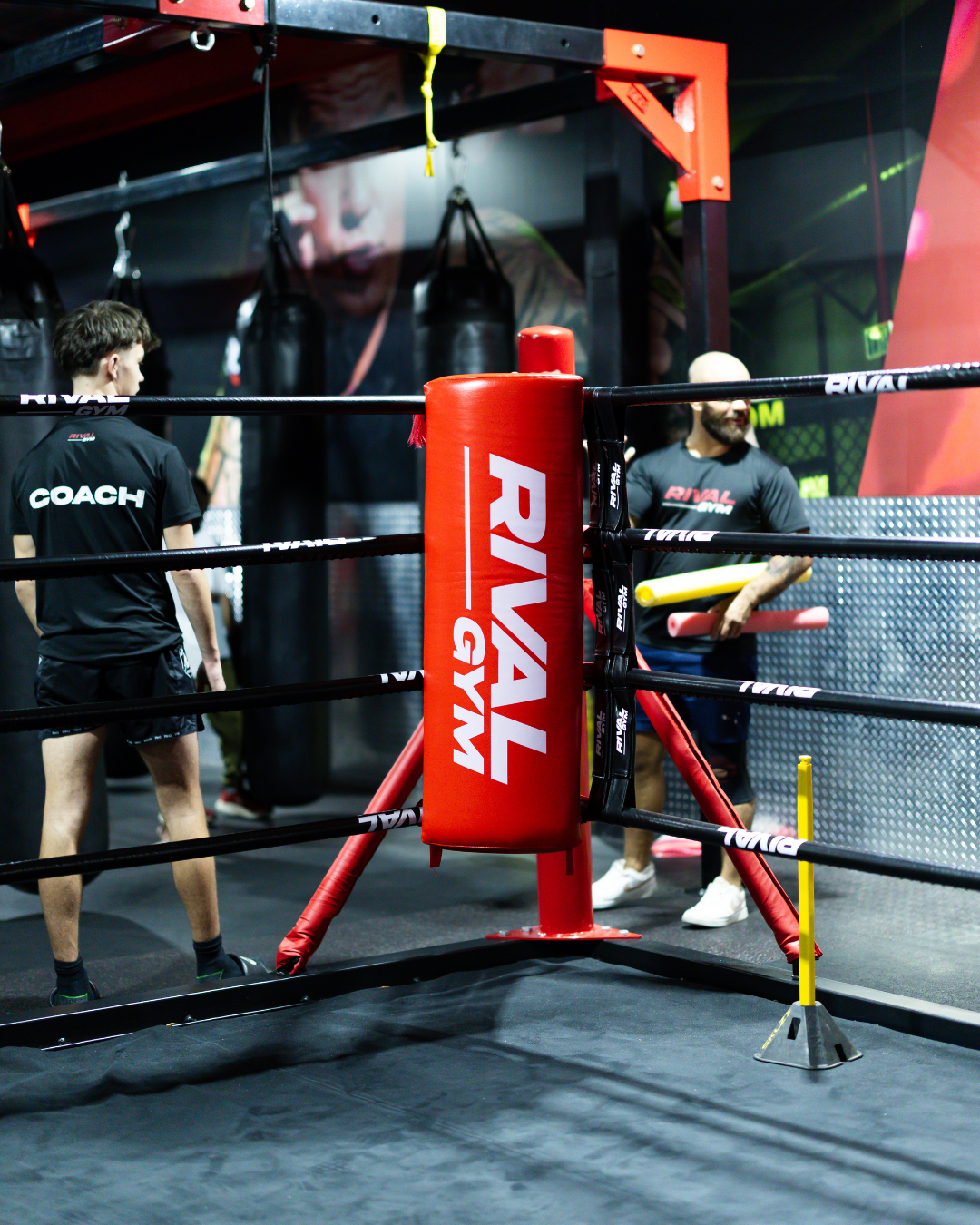 Inside a boxing gym with a boxing ring, a coach, and a trainer holding foam sticks. There are punching bags in the background and a large poster of a boxer on the wall.
