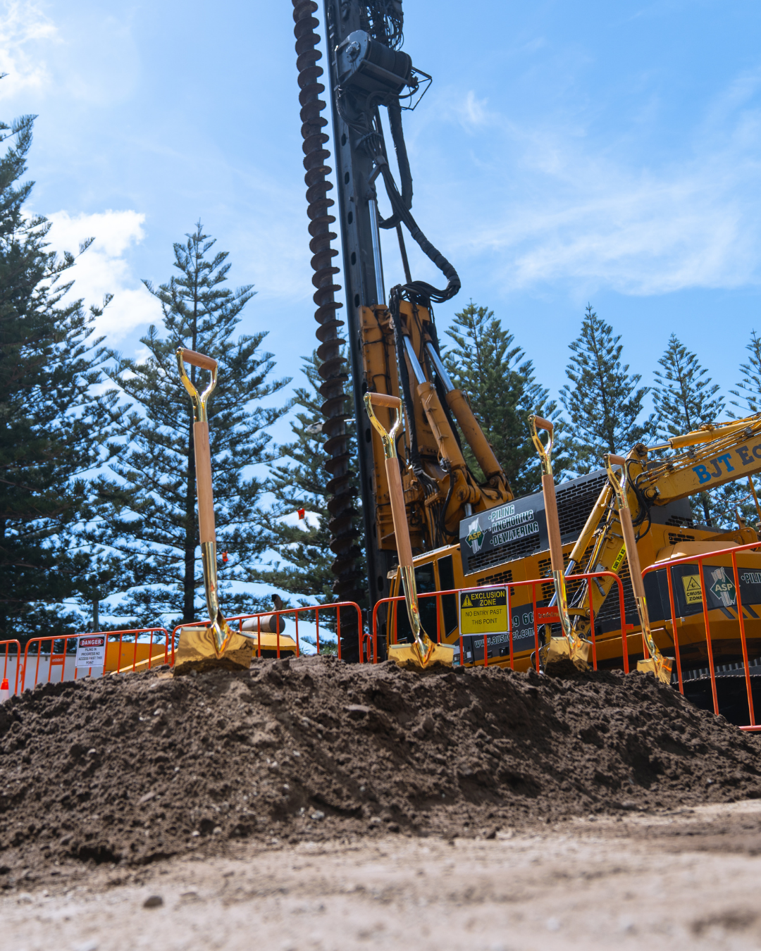 A large construction drill machine on a construction site with dirt in the foreground, orange safety barriers, and tall pine trees in the background under a blue sky.