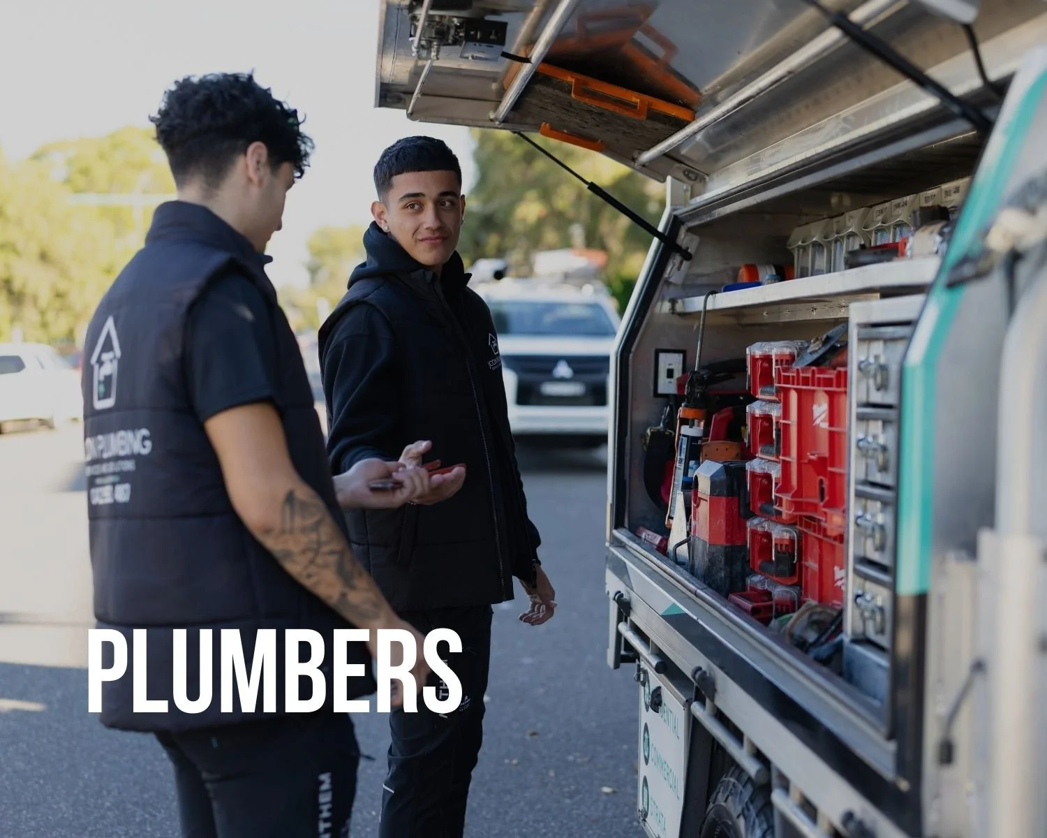 Two young male plumbers standing outdoors next to a mobile tool cart with various tools and equipment, having a conversation.