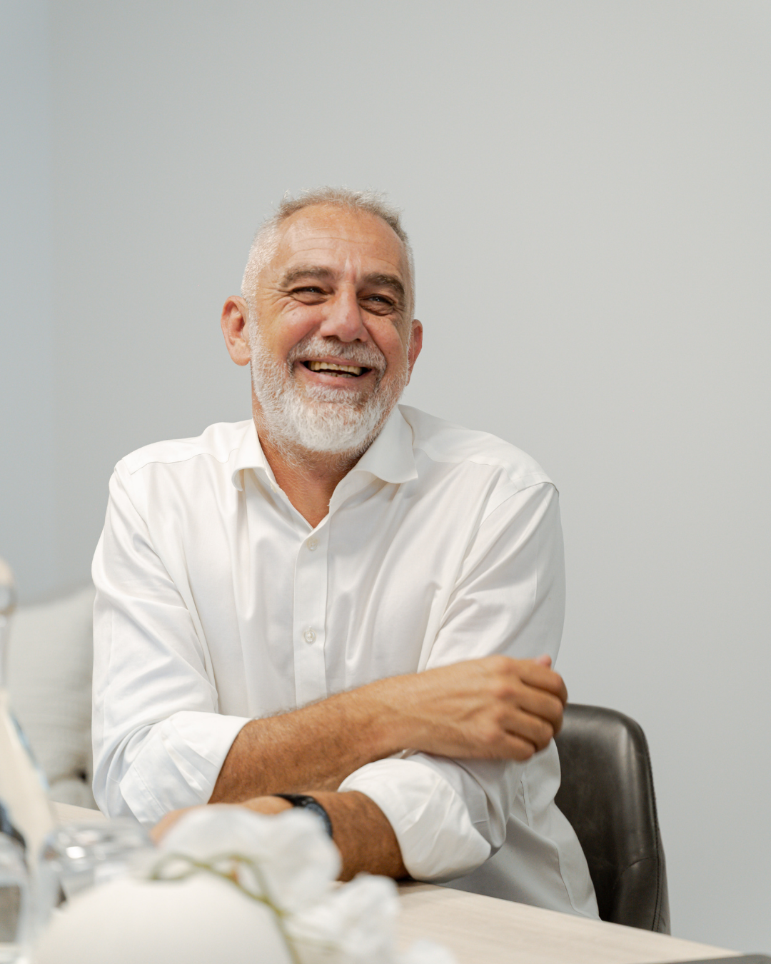 A smiling older man with gray hair and beard wearing a white shirt sitting at a table.