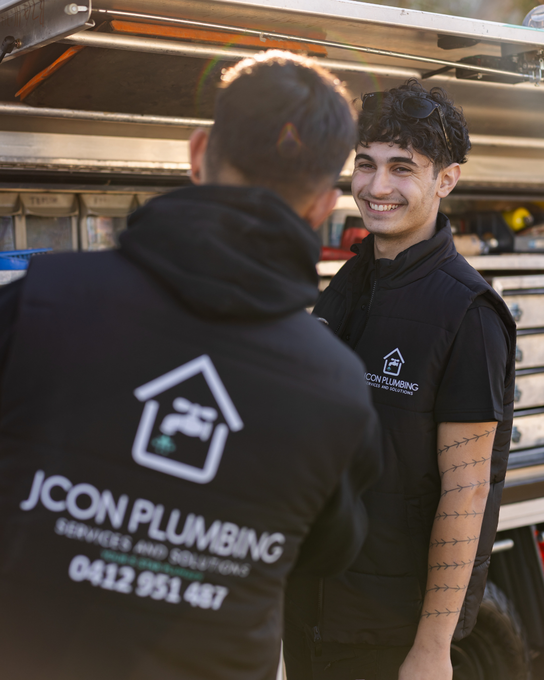 Two HVAC technicians from JCON Plumbing smiling and talking outdoors near a service truck with tools.