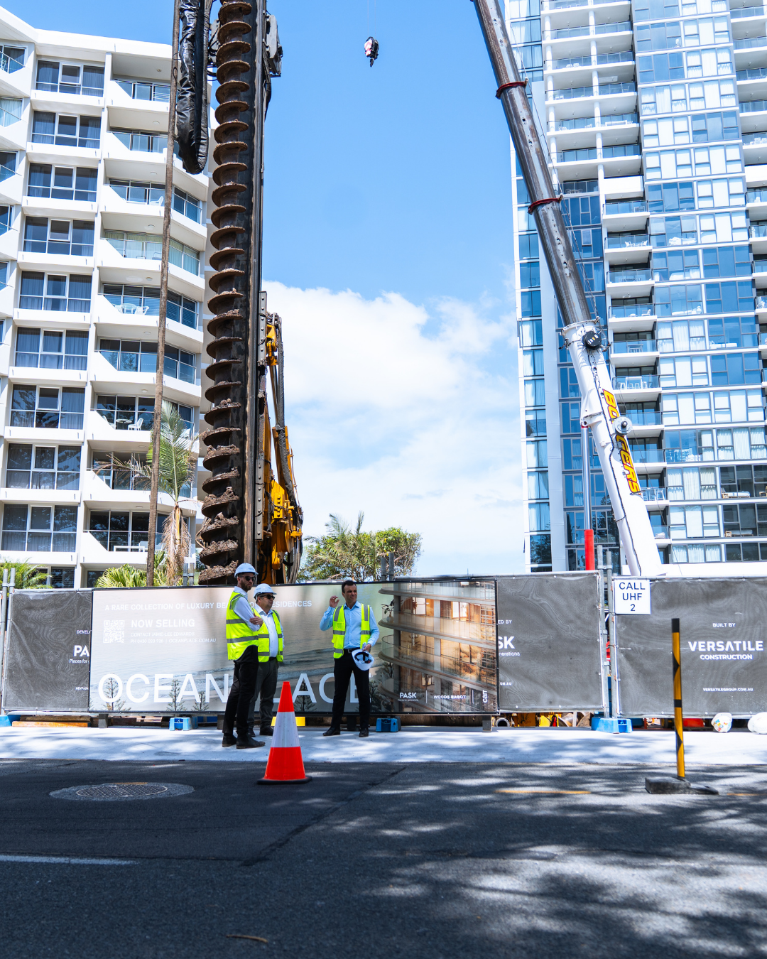 Construction workers in high-visibility vests and hard hats standing near large construction equipment at a building site between modern high-rise apartment buildings on a sunny day.