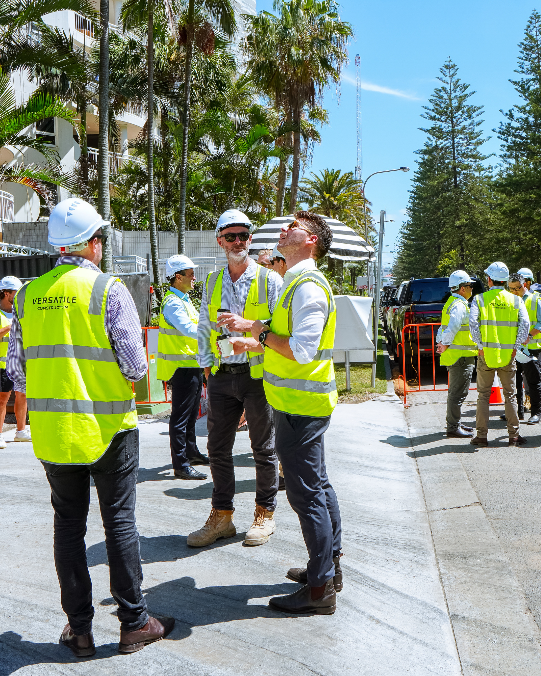 Construction workers in high-visibility vests and hard hats having a discussion on a sunny day, with palm trees and a building in the background.