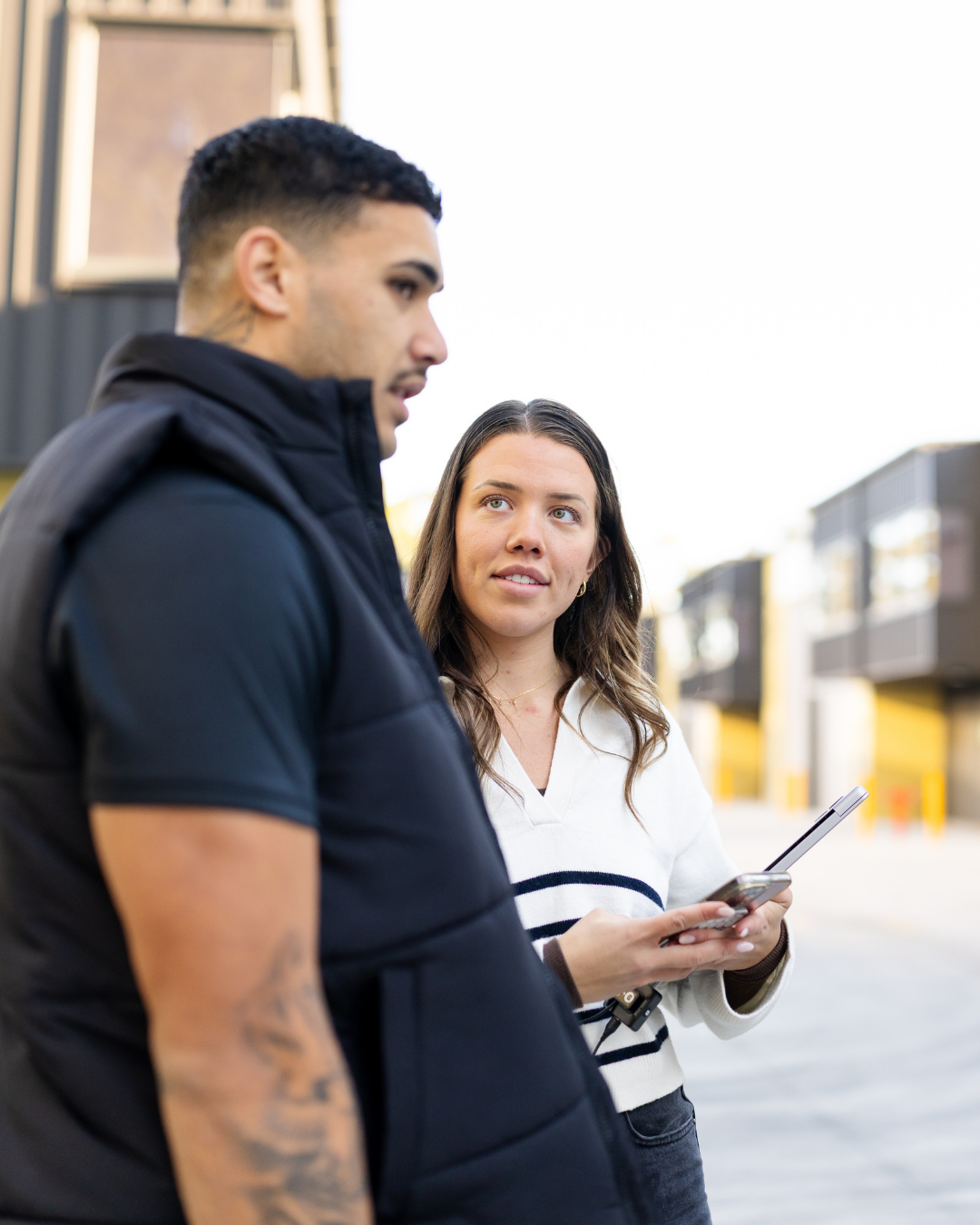A woman holding a smartphone looks at a man standing beside her outdoors in a commercial area with building overhangs.