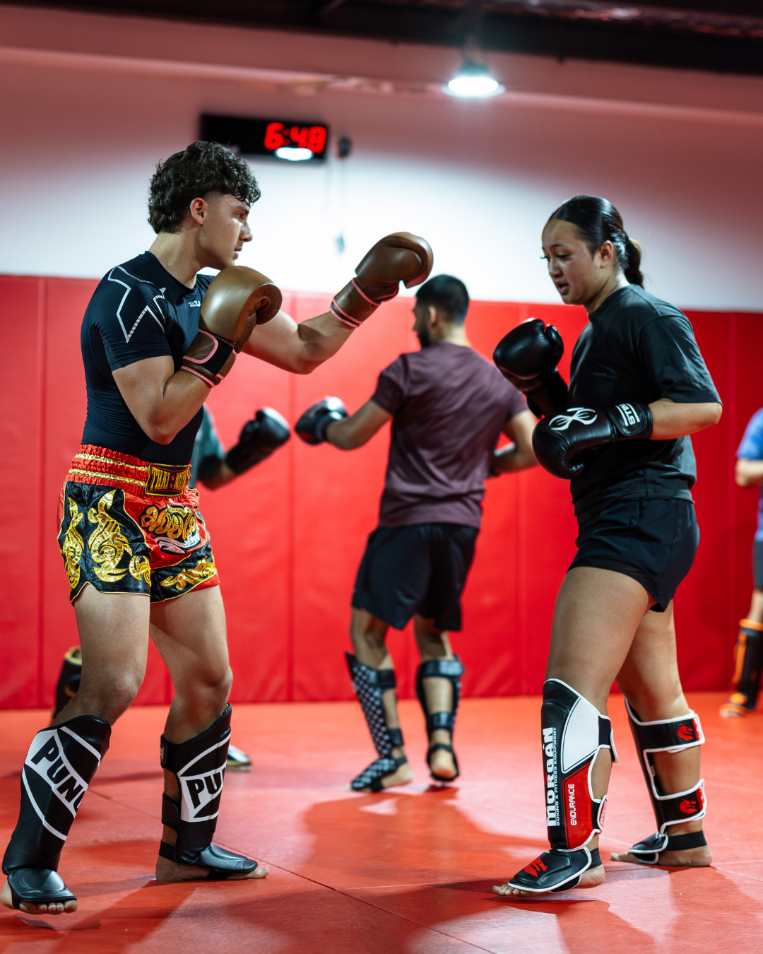 Two female Muay Thai fighters practicing with gloves and shin guards in a gym, with two male fighters in the background.