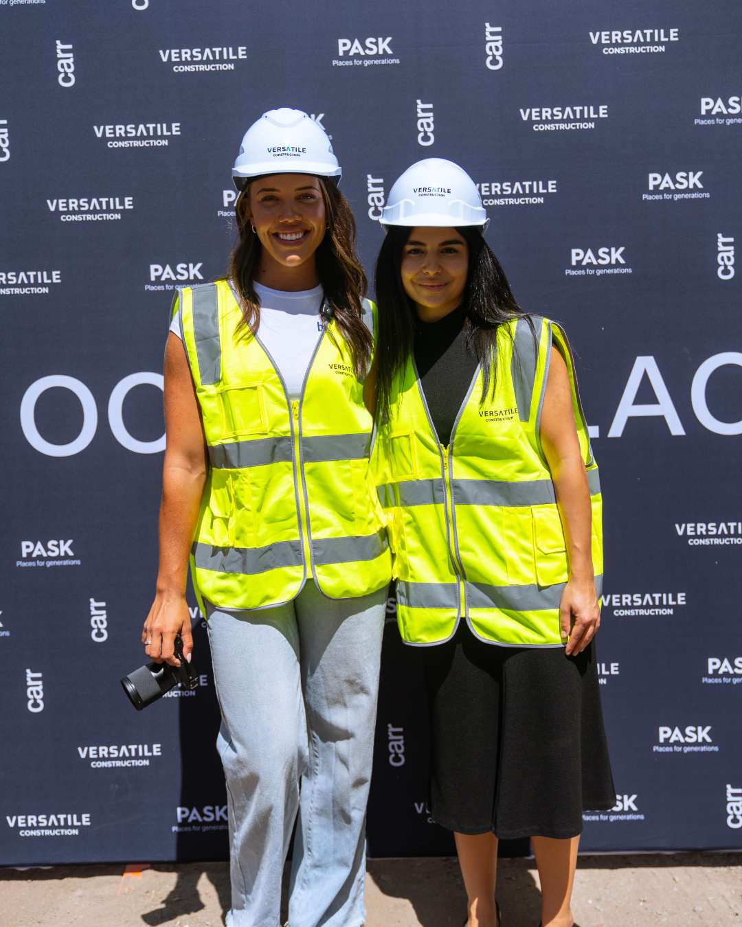 Two women wearing yellow safety vests and white construction helmets standing outdoors, smiling for the camera in front of a dark backdrop with company logos.