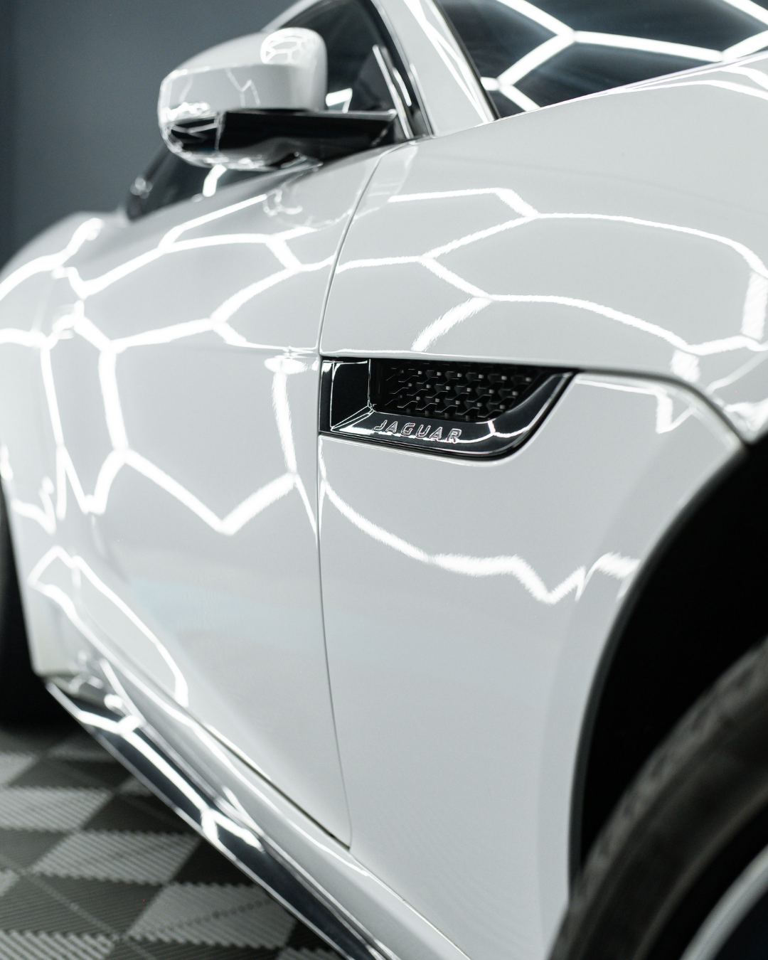 Close-up of a white Jaguar car showing the front fender and side mirror with reflections of linear lights.