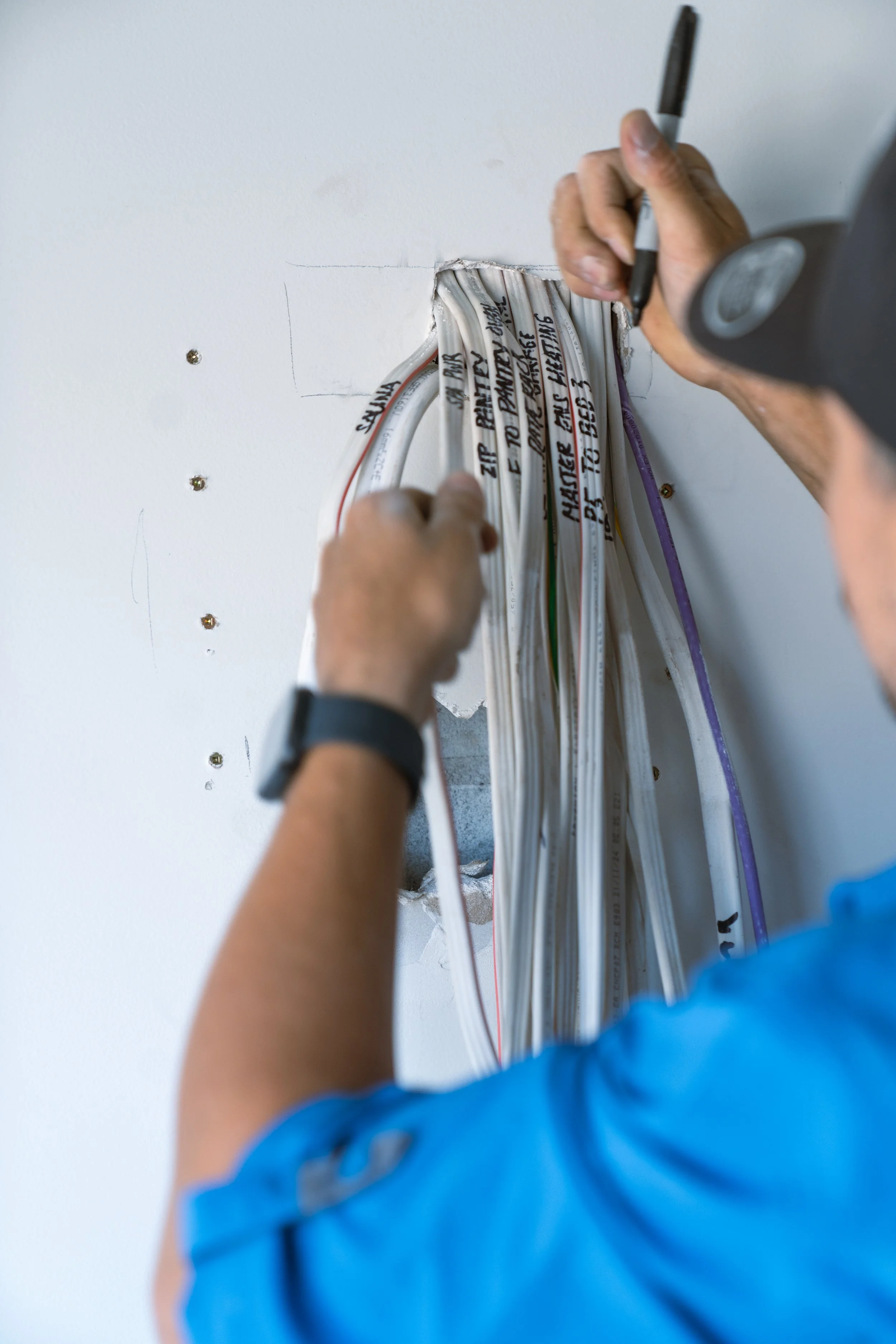A technician in a blue shirt working on electrical wiring in a wall, marking and organizing cables.