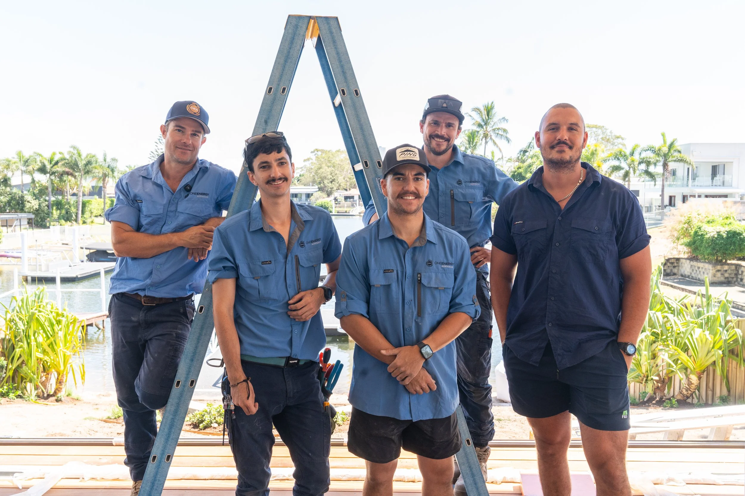 Six men standing outdoors near a marina with boats and palm trees, some wearing blue shirts with a logo and one in a dark shirt and shorts, smiling for the photo with a blue ladder in the background.