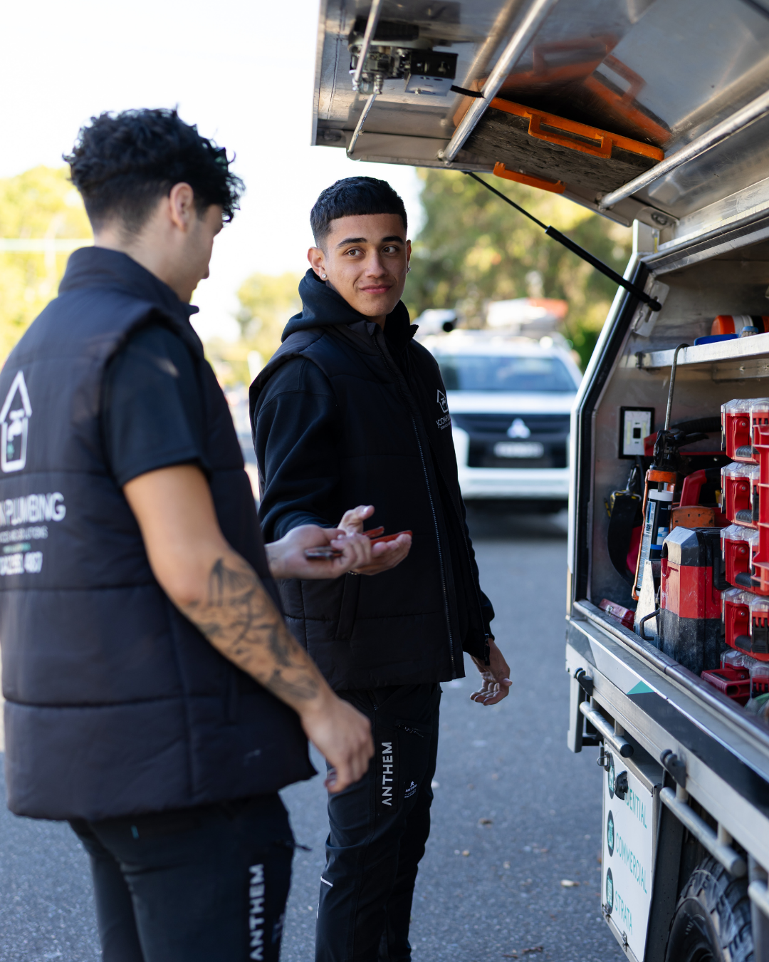 Two young men in black jackets talking beside a mobile work van, which has tools and equipment stored in its open compartments, on a city street during daytime.