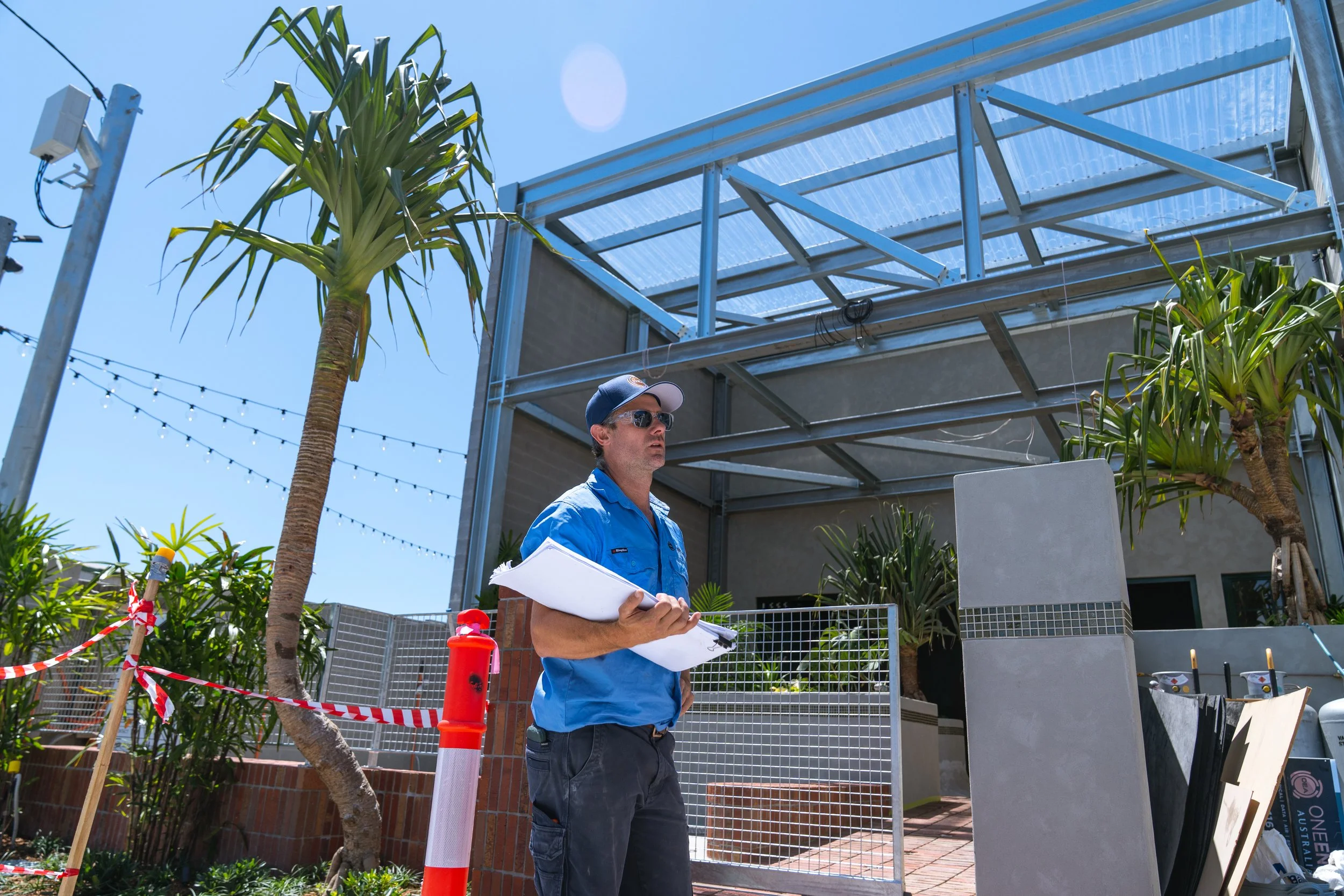 A man in sunglasses and a blue shirt holding papers, standing outside a construction site near some palm trees and a partially built structure with a metal framework.