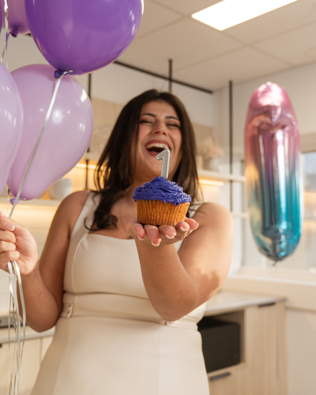 A woman with dark hair celebrating her 1st birthday, holding a cupcake with purple frosting and a number 1 candle, smiling and with balloons around her in a decorated indoor setting.