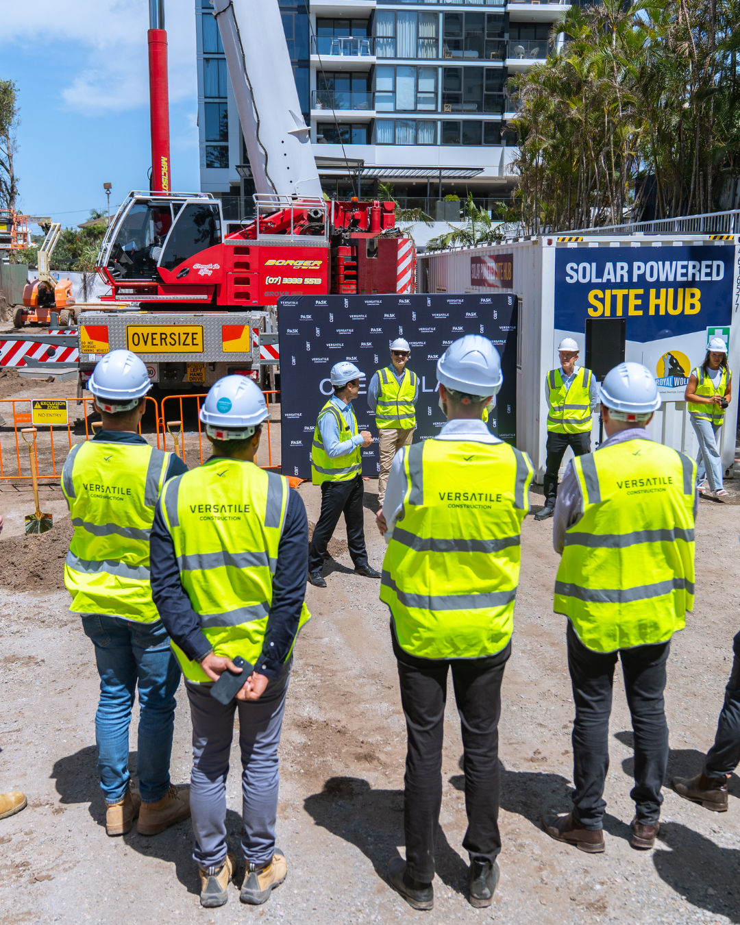 Group of construction workers and professionals in yellow safety vests and white hard hats gathered at a construction site, with a large red crane and a blue and white sign that reads 'SOLAR POWERED SITE HUB' in the background.