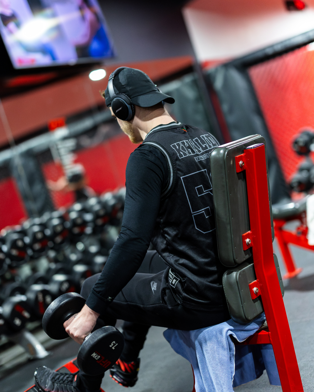 A man performing a seated bicep curl exercise at the gym, wearing headphones, a black cap, black long-sleeve shirt, and black pants, with dumbbells in his hands, in a gym with red and black equipment.