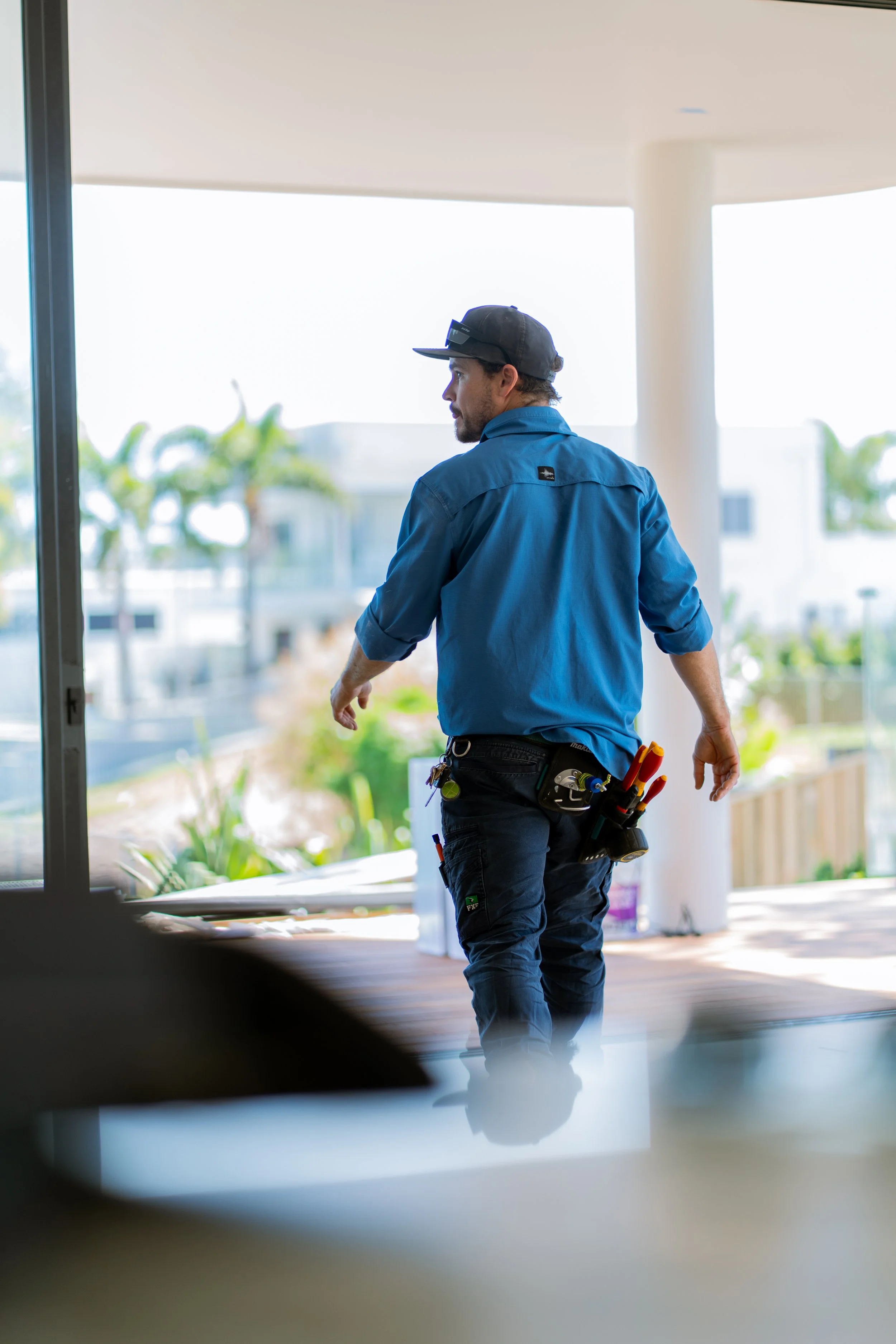 Man in blue work shirt and black cap walking indoors, tool belt with tools around his waist, sunlight coming through large windows, plants outside visible.