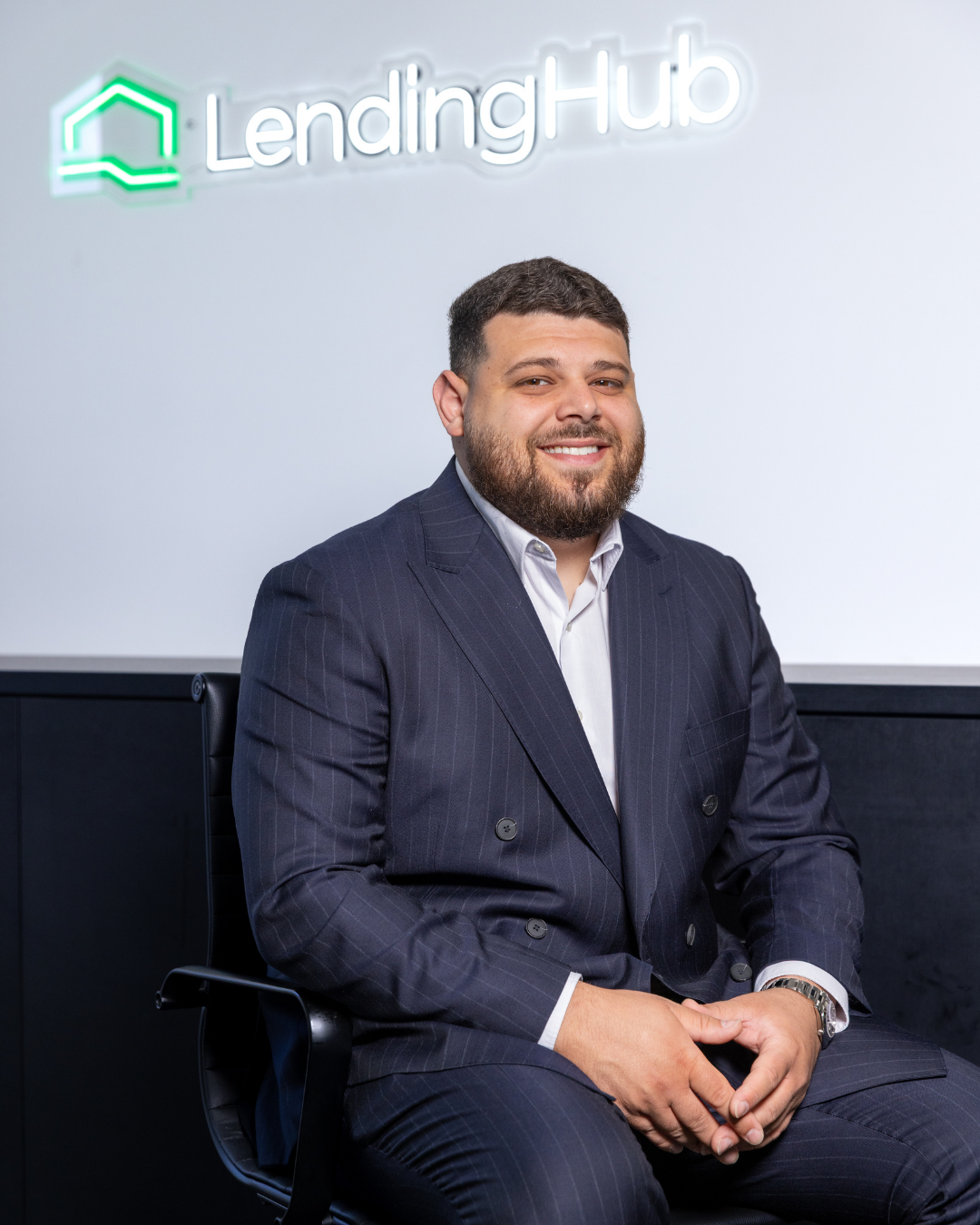 Man in a suit sitting in front of a lit sign that reads 'LendingHub' with a house logo.