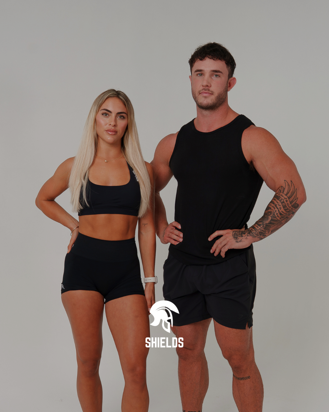 Two fit individuals, a woman and a man, standing side by side in athletic wear against a plain white background. The woman has long blonde hair, is wearing a black sports bra and matching shorts, and has her right hand on her hip. The man has short d