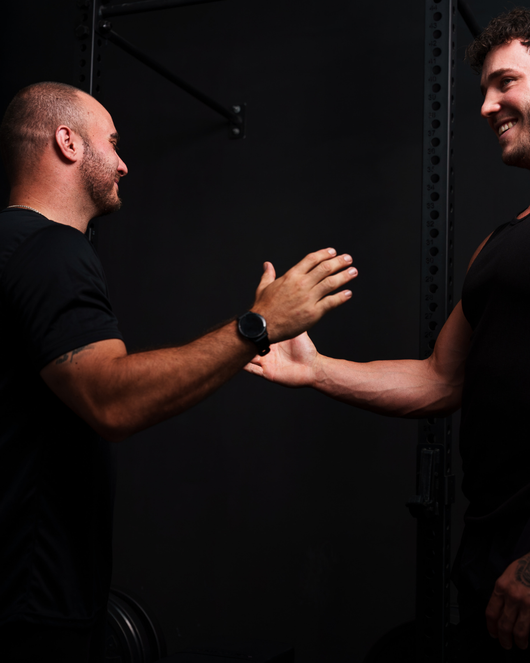 Two men in sports apparel greeting each other with a handshake and smile in a gym.