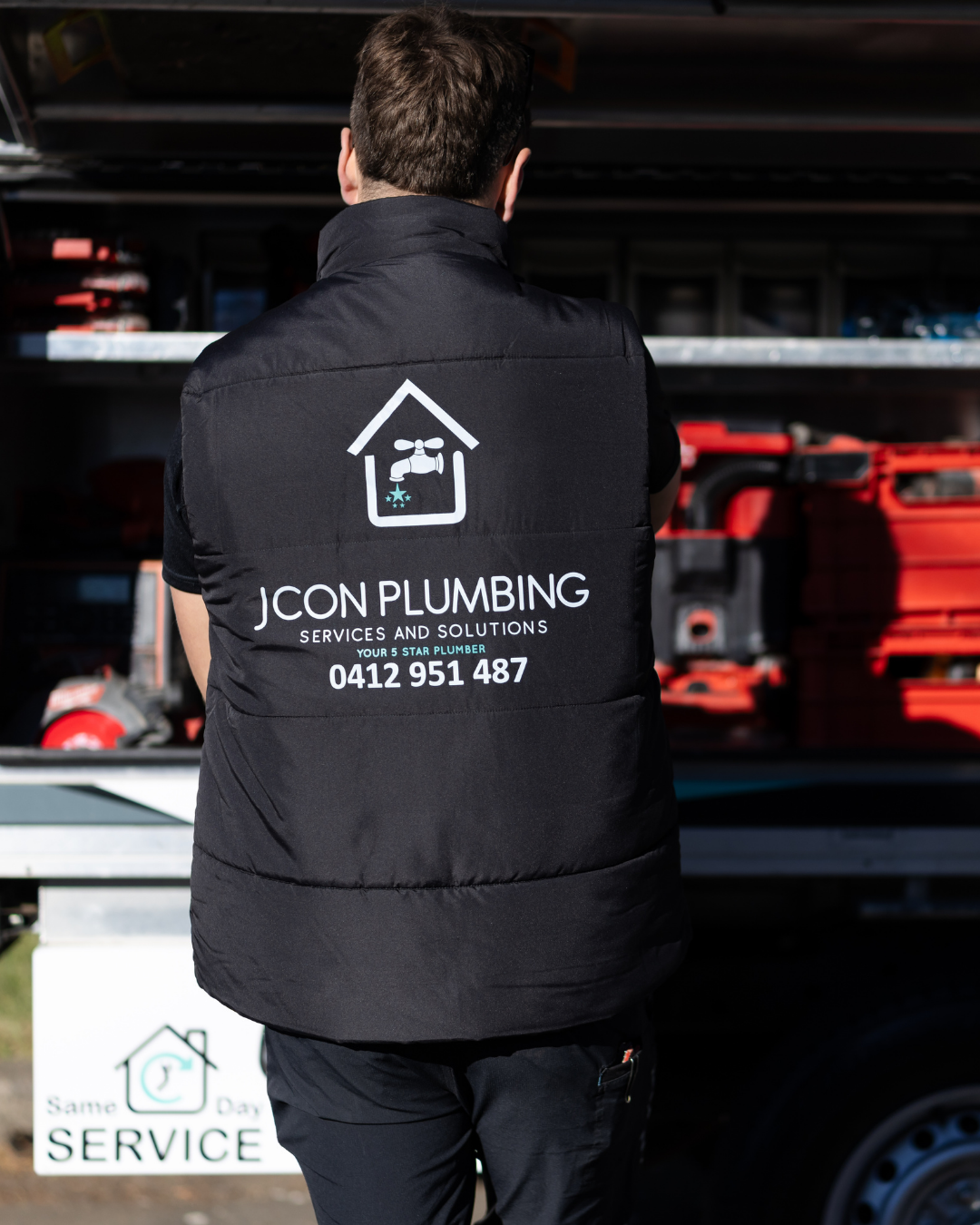 A person standing with back to the camera, wearing a black vest with logo and contact information for JCON Plumbing, against a background of tools and equipment on store shelves.