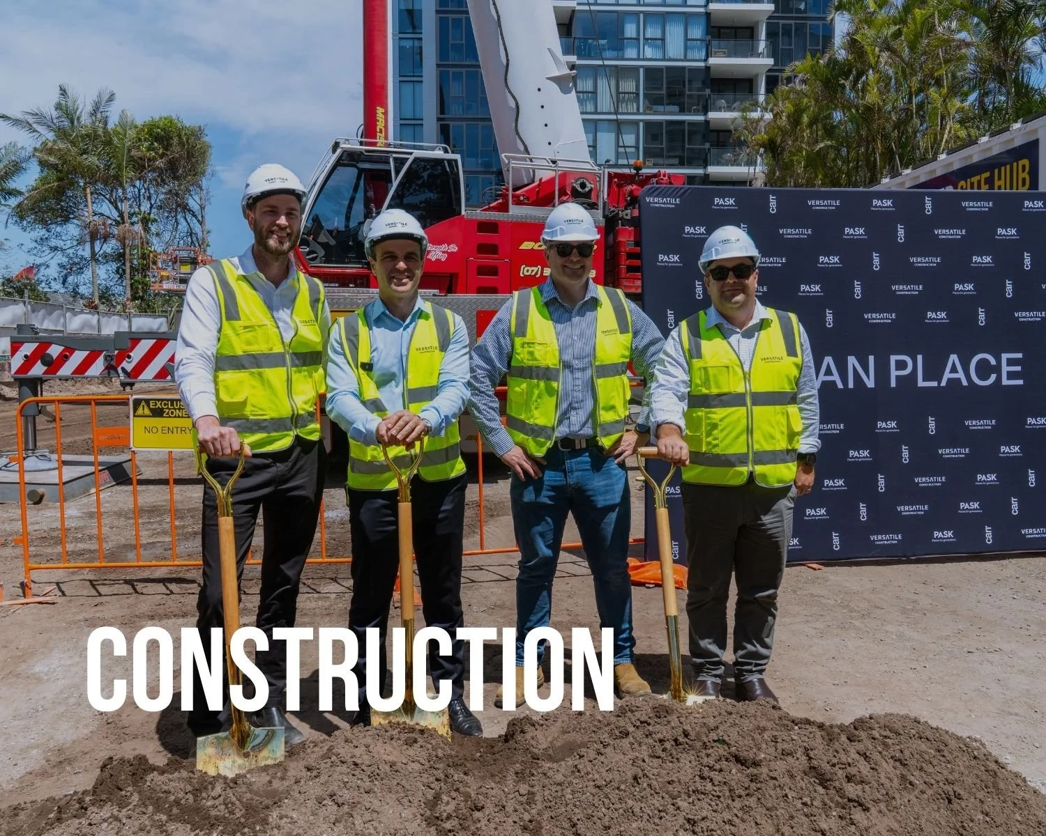 Four men in construction vests and helmets holding shovels at a groundbreaking event on a construction site with a red crane and high-rise building in the background.