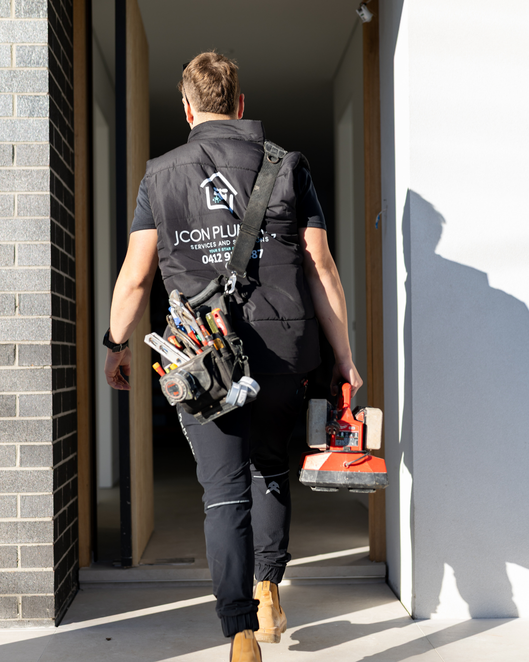 A handyman carrying a tool bag filled with various tools and a power saw, walking into a building.