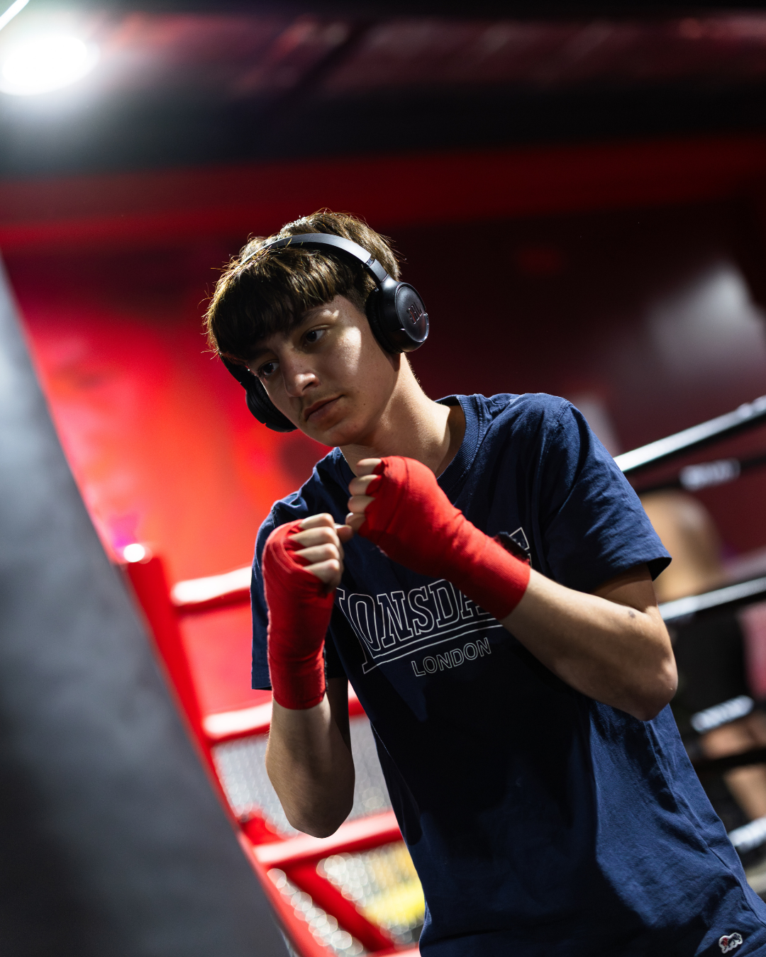 A young male boxer with red hand wraps in a boxing gym, wearing headphones and in a fighting stance.