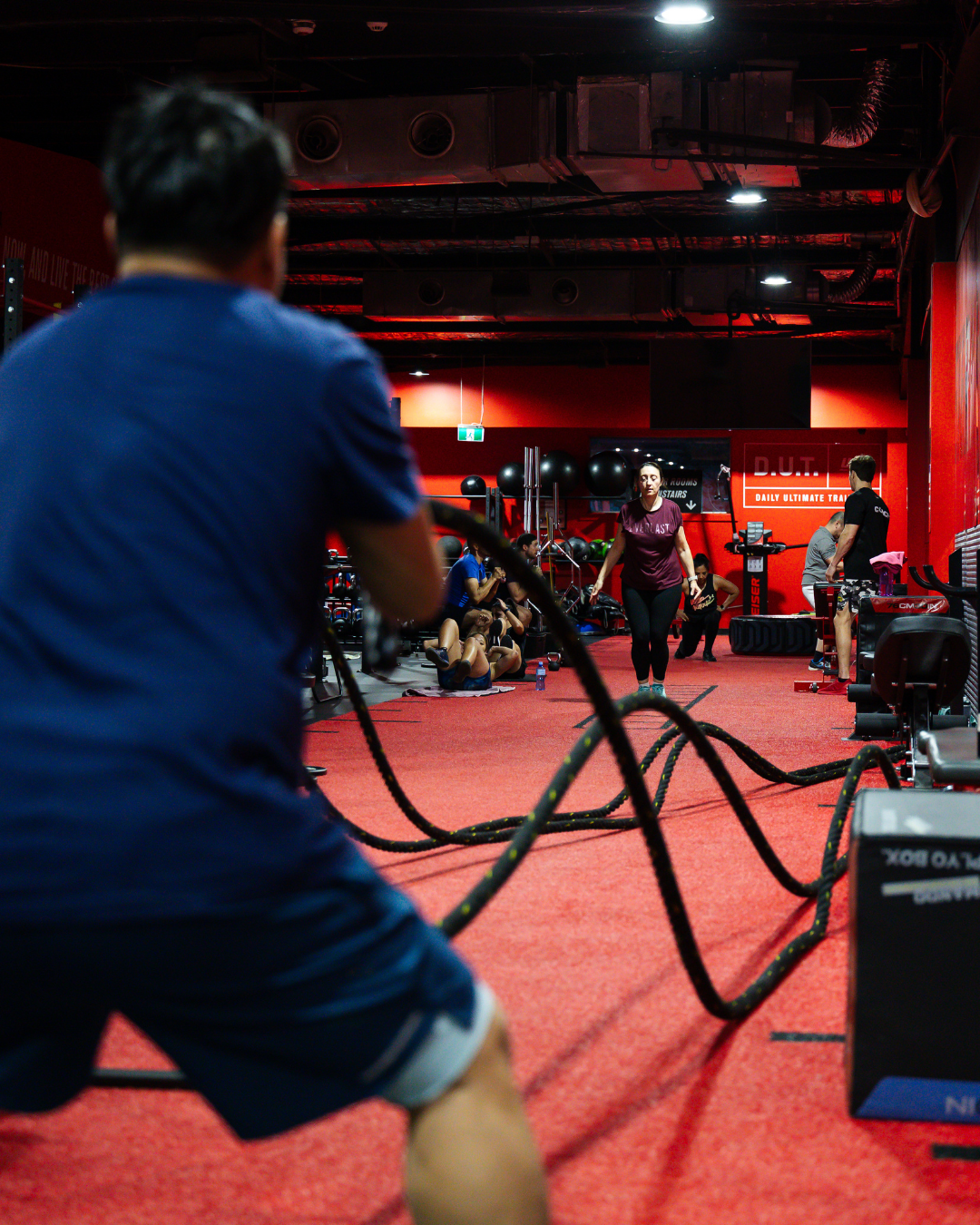 People participating in a workout class using battle ropes in a gym with red walls, fitness equipment, and a group exercise session.