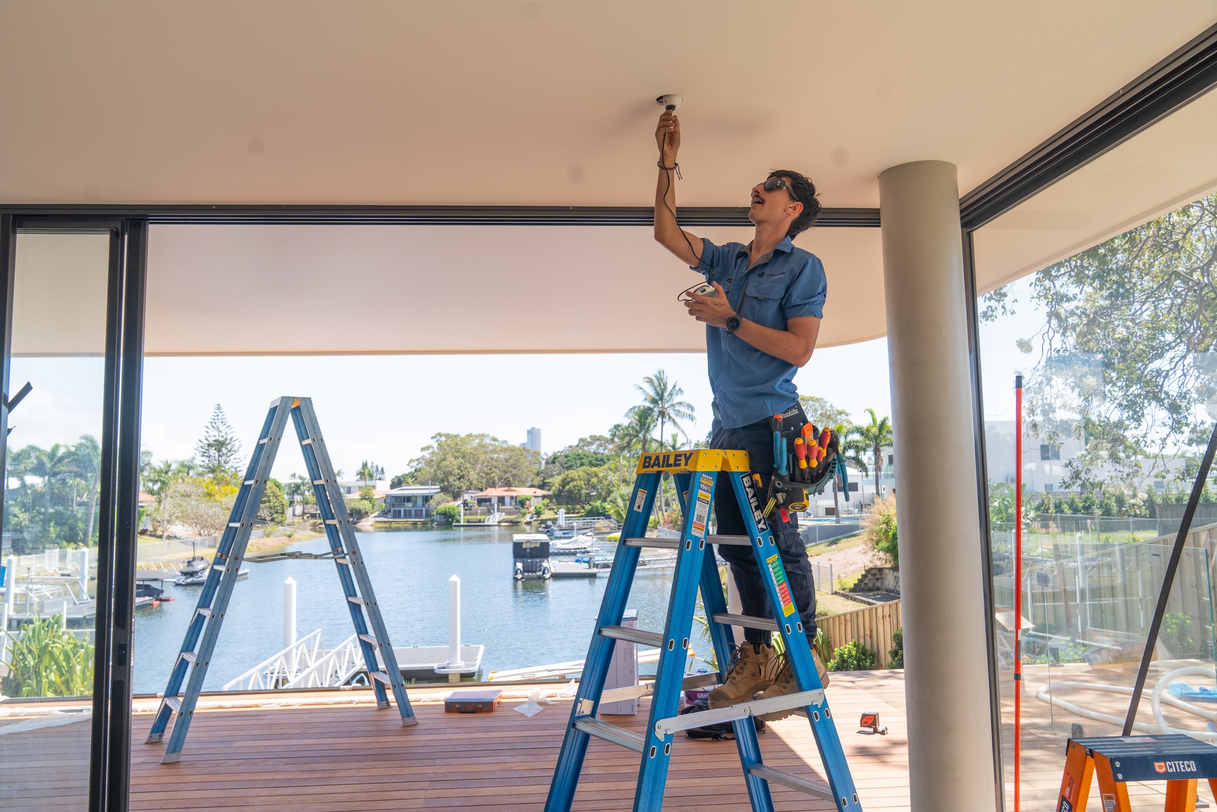 A man standing on a ladder installing a ceiling light fixture in a modern room with large windows overlooking a waterway with boats and houses, with tools attached to his belt.