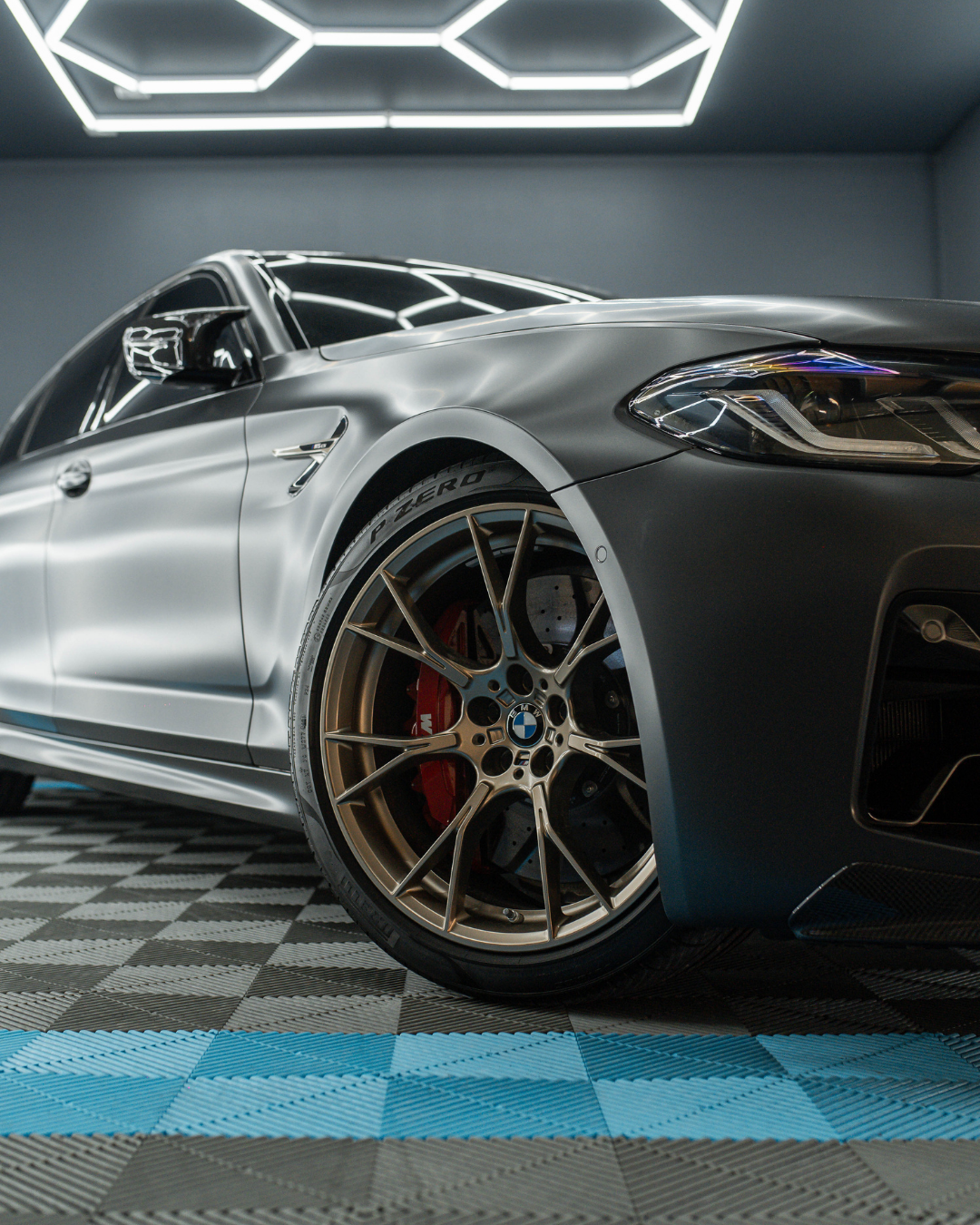 Close-up of a silver BMW sports car showcasing the front right wheel and headlight, in a modern garage with geometric ceiling lighting.