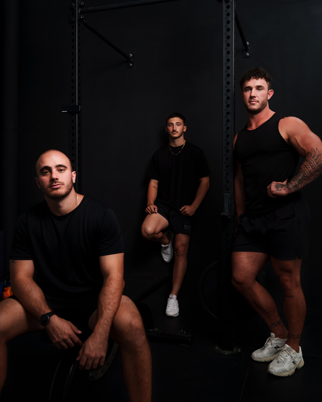 Three young men in black athletic clothing in a gym against a black wall, one sitting, one leaning on a bench, and one standing, all looking at the camera.