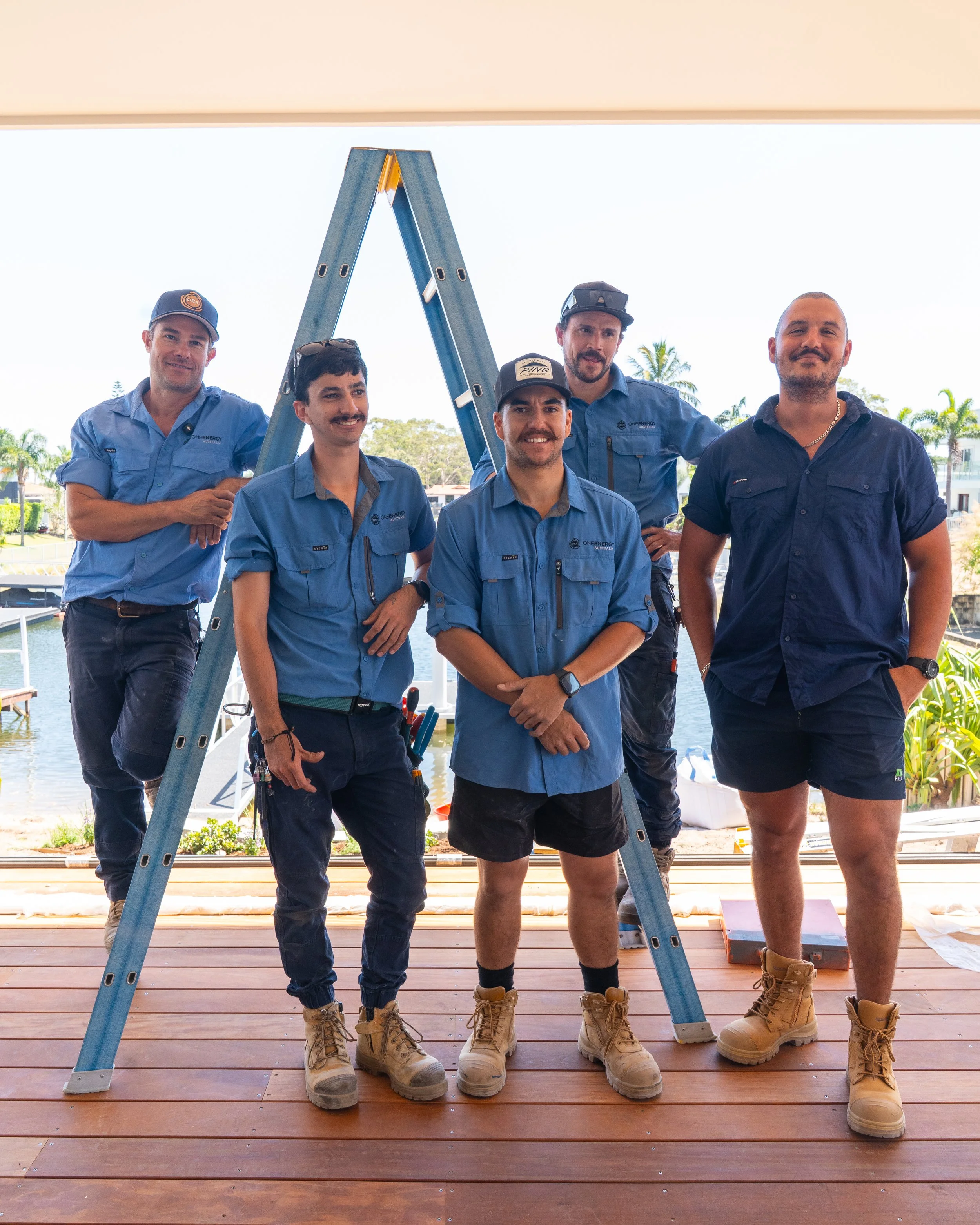 Five men in work shirts and boots standing on a wooden deck with a view of water and palm trees in the background, posing for a photo.