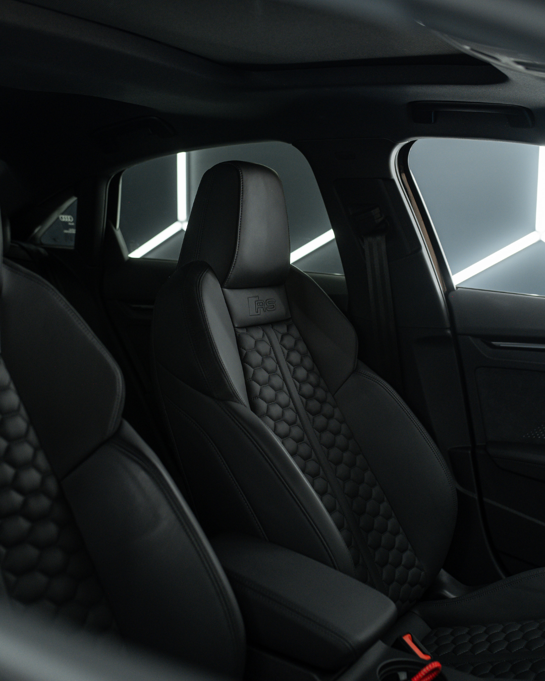 Close-up of a black car interior showing a leather racing seat with honeycomb pattern and RS logo, part of the front passenger area.