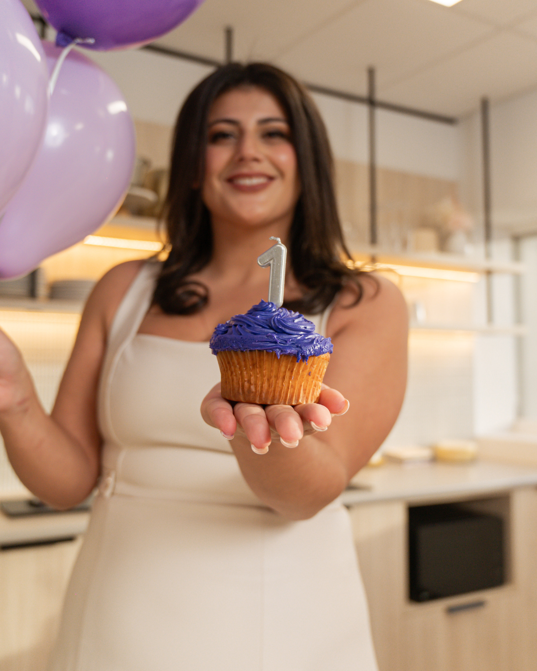 A woman holding a cupcake with purple frosting and a '1' candle, celebrating her first birthday.