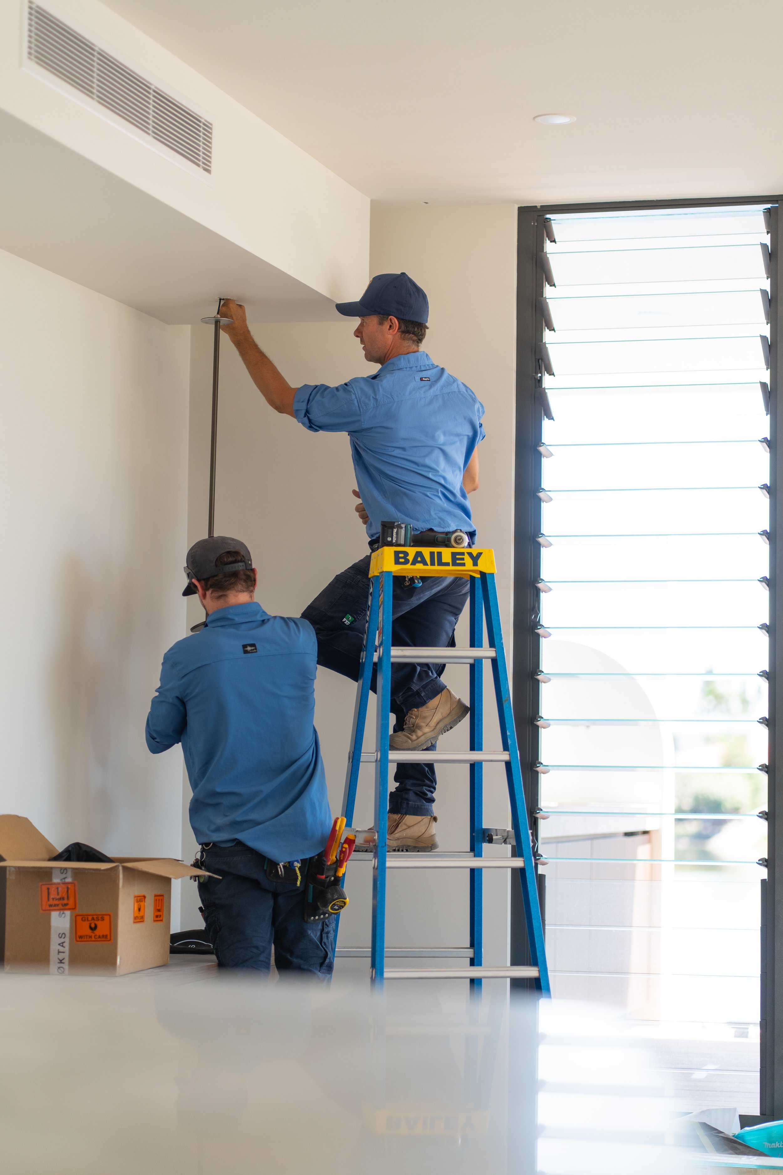 Two workers installing or repairing a ceiling vent or air conditioning unit, one standing on a ladder labeled 'BAILEY' and the other on the ground assisting, inside a room with window louvers.