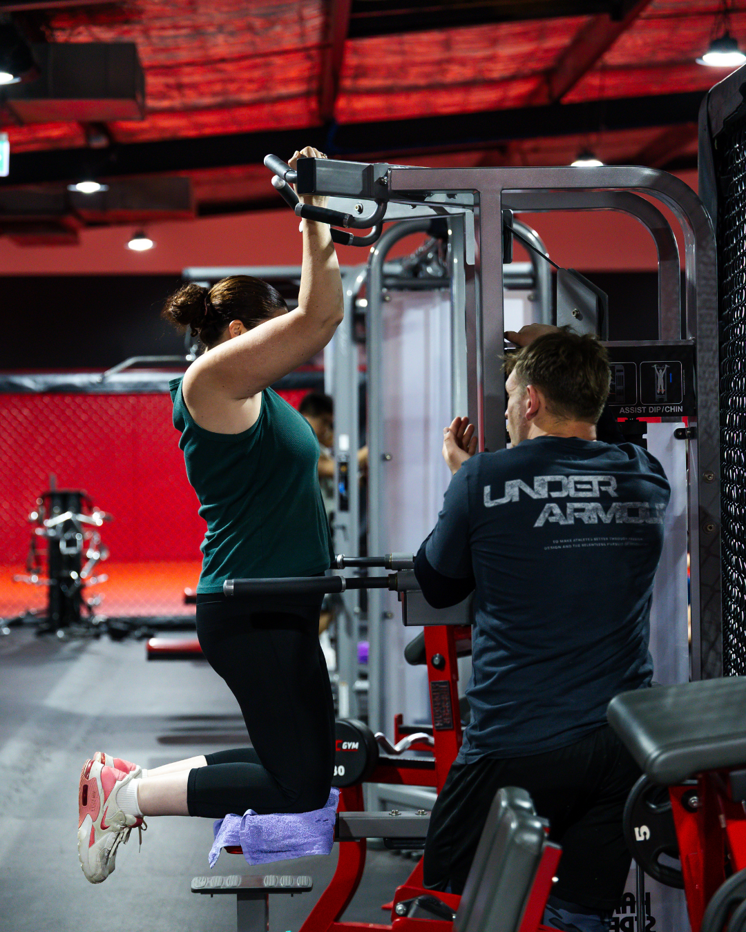 A woman performing tricep dips on equipment while a trainer assists her at a gym.