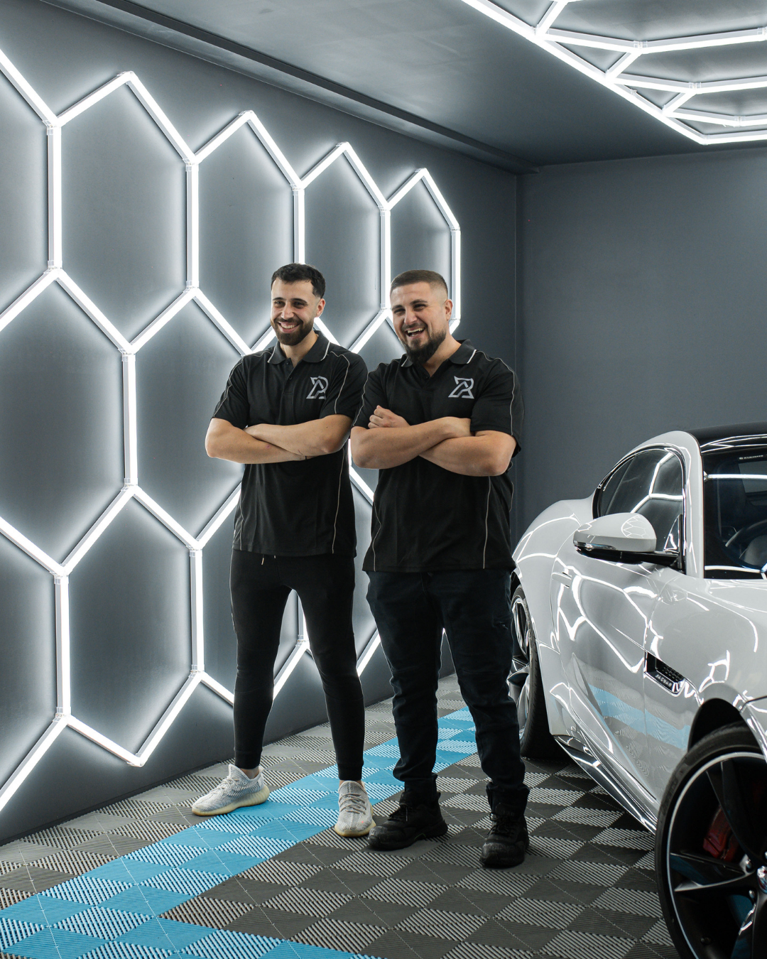 Two men standing inside a modern garage with a white sports car; both are smiling, crossing their arms, and wearing black polo shirts with a logo, in front of a wall with geometric neon lighting.