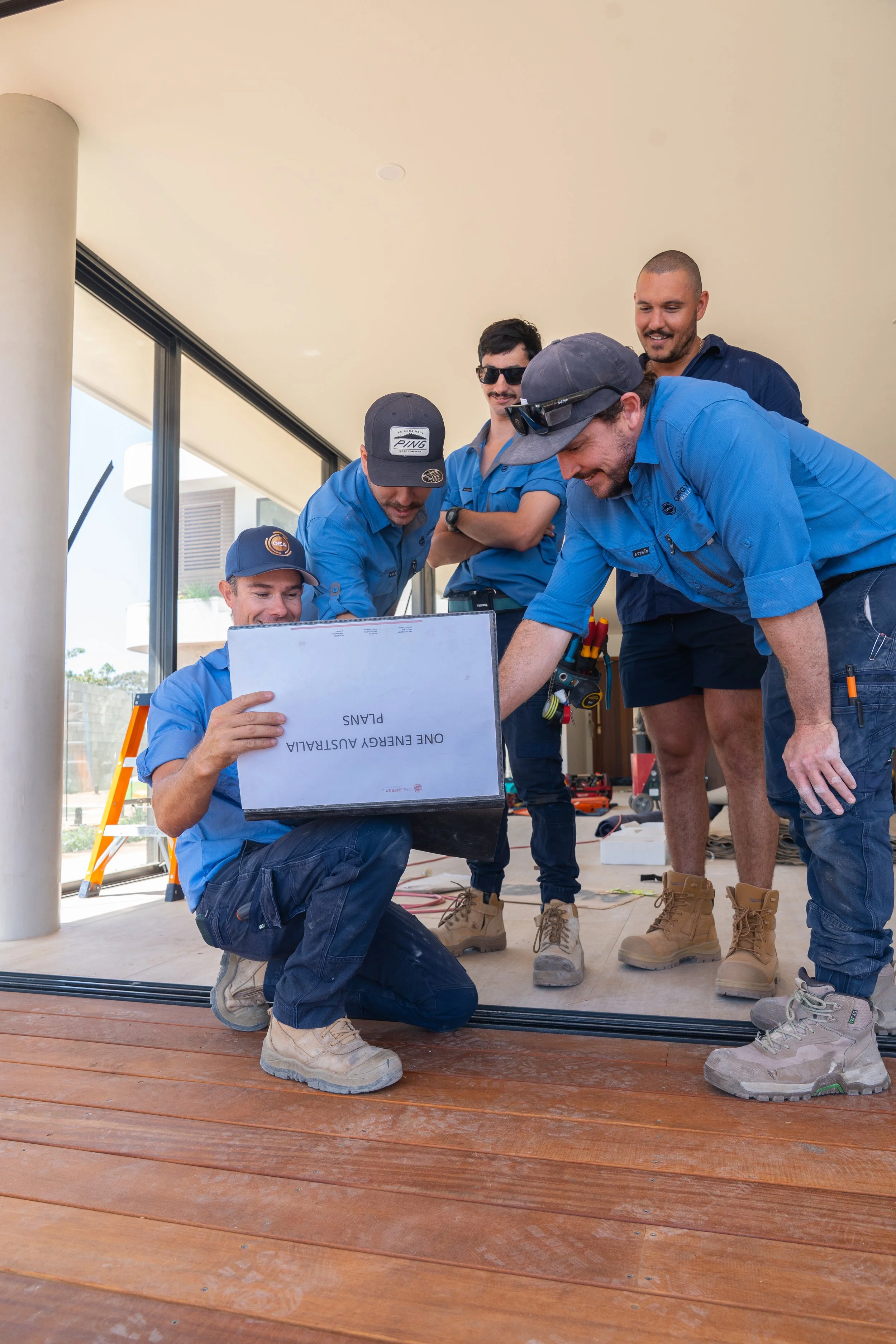 Group of construction workers wearing blue uniforms and boots gathered around looking at a laptop, inside a building with large windows and wooden floors.