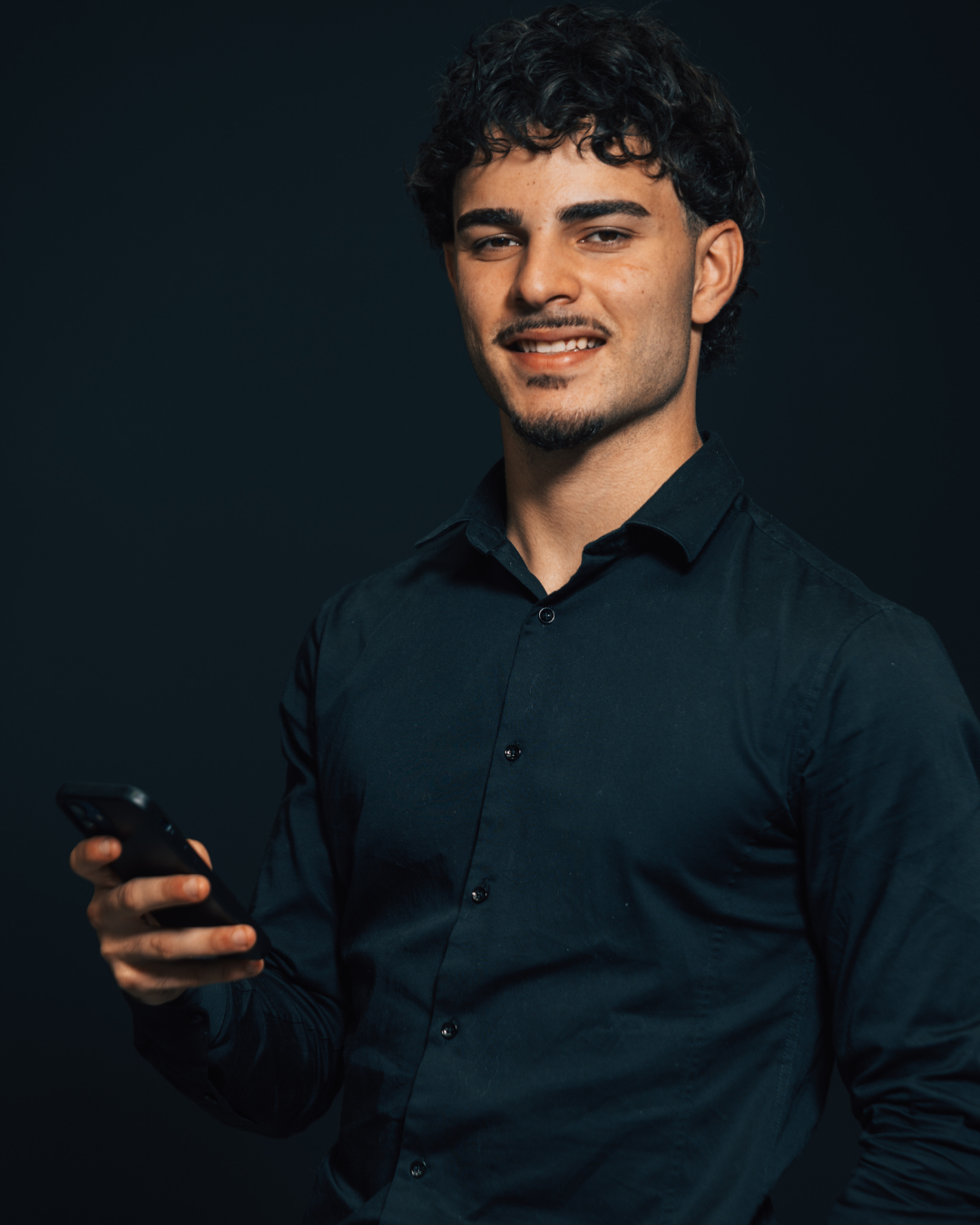 A young man with dark curly hair and a goatee, wearing a black shirt, holding a smartphone, standing against a dark background.