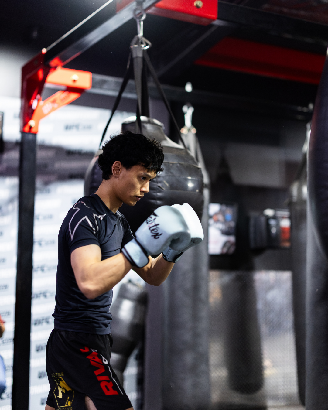 A young male boxer practicing in a gym, wearing boxing gloves and shorts, standing near a punching bag.